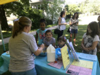 A group of children and adults gather around a table at an outdoor event, where a woman is serving refreshments to kids, with colorful signs and a milk jug visible on the table.