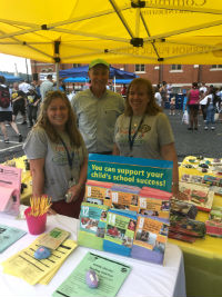 Three individuals stand behind a table filled with informational materials and school supplies, under a yellow canopy at an outdoor event.