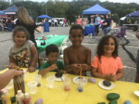 Four children are sitting at a table covered with a yellow cloth, engaged in a painting activity with various colors and materials, while a crowd and tents are visible in the background.