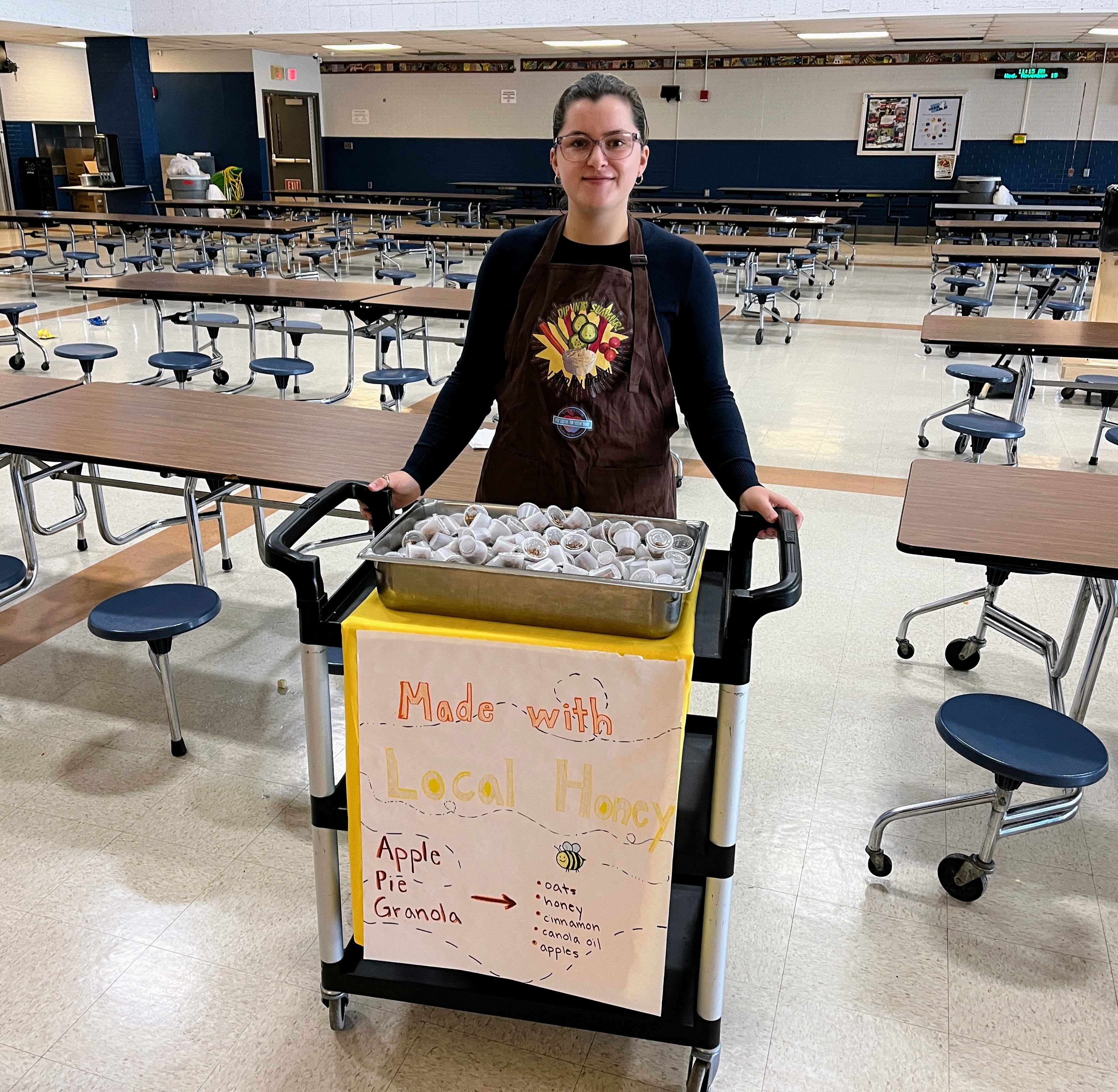 A person stands behind a cart filled with food, displaying a sign that reads 'Made with Local Produce' in a cafeteria setting with empty tables in the background.