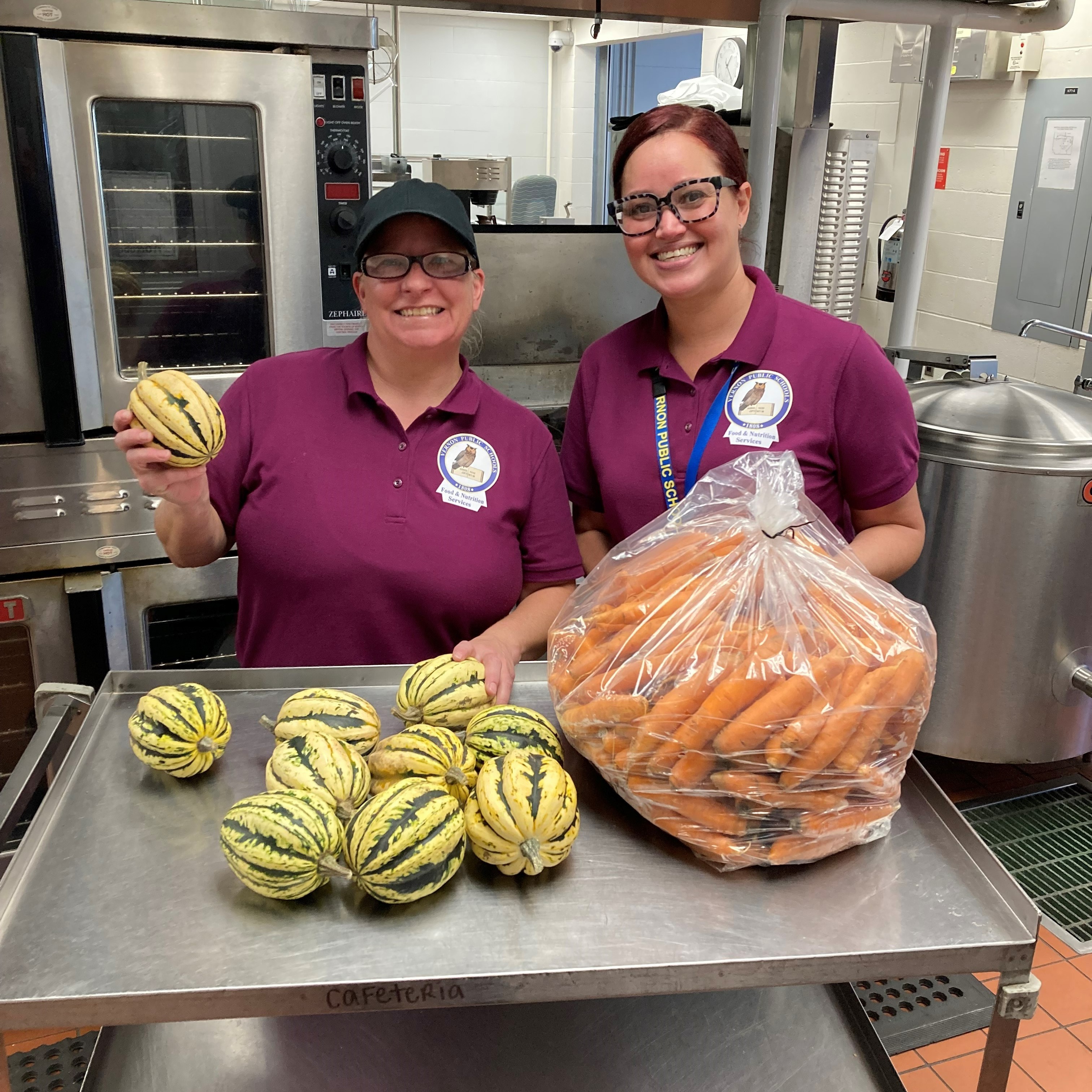 Two women in purple shirts smile while holding squash and a bag of carrots in a kitchen setting.