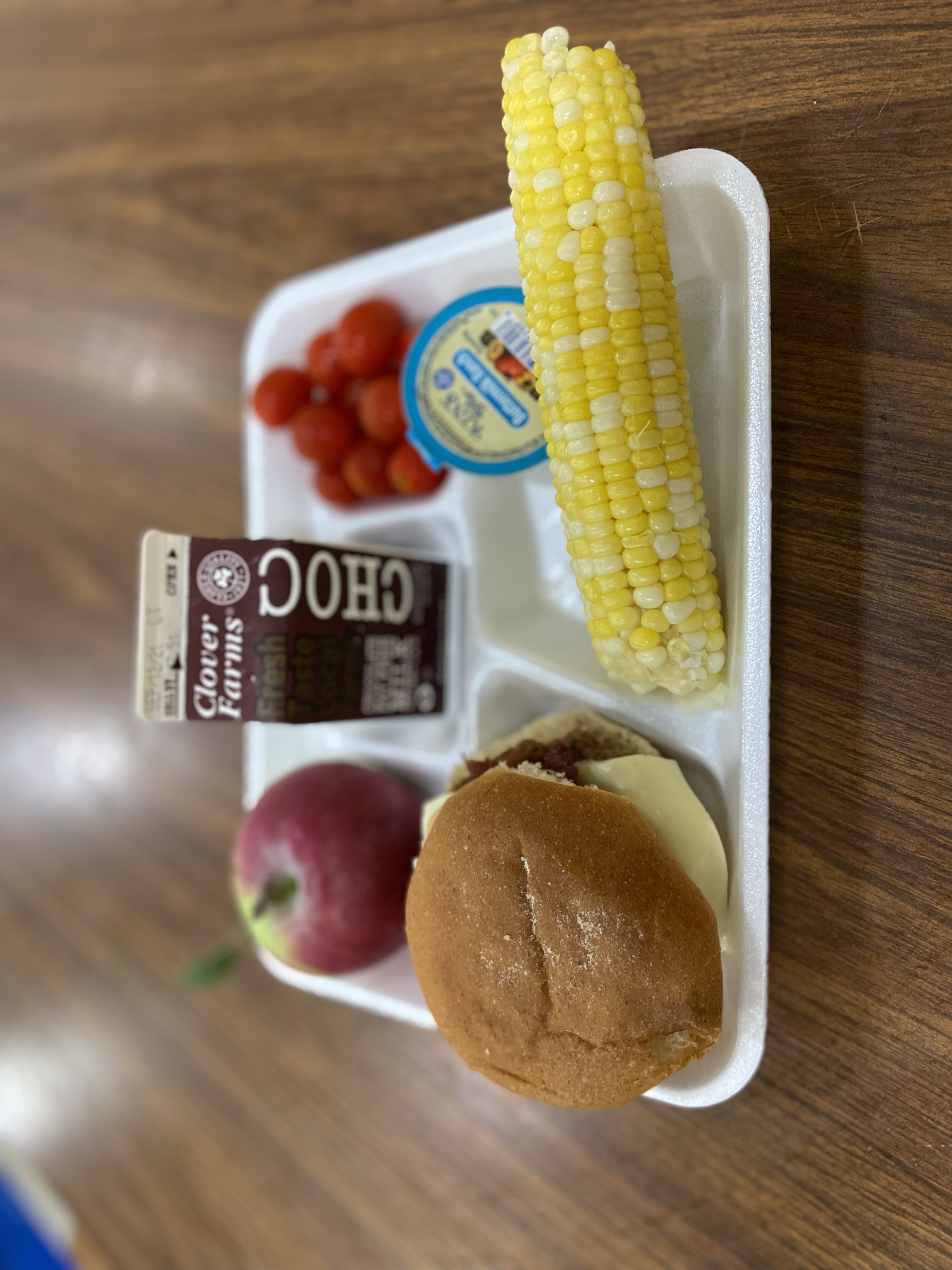 A school lunch tray featuring a hamburger, a carton of chocolate milk, an apple, cherry tomatoes, a serving of corn on the cob, and a small container of dipping sauce.