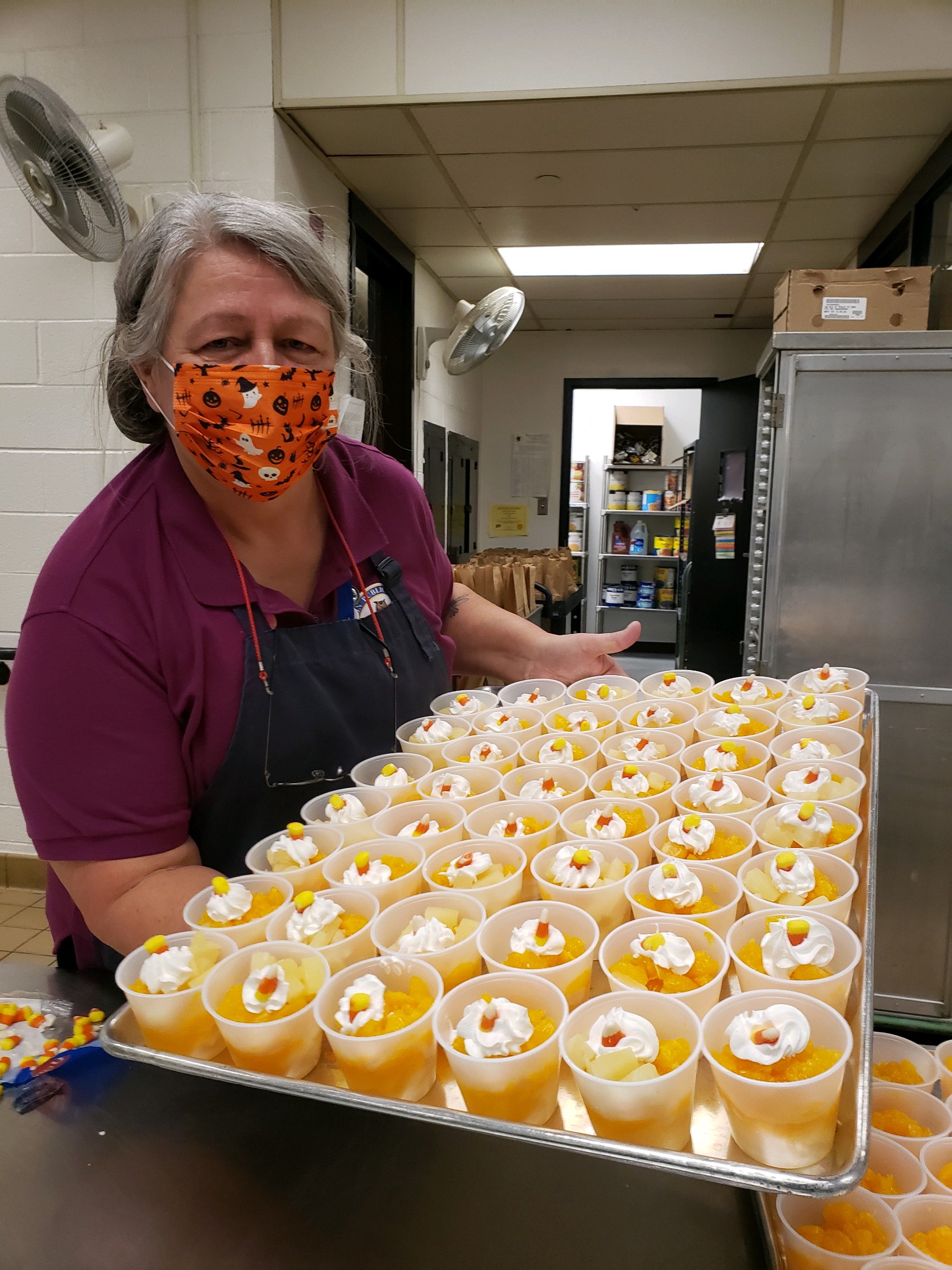 A woman wearing a colorful mask smiles while holding a tray filled with numerous cups of dessert topped with whipped cream and fruit, set in a kitchen environment.