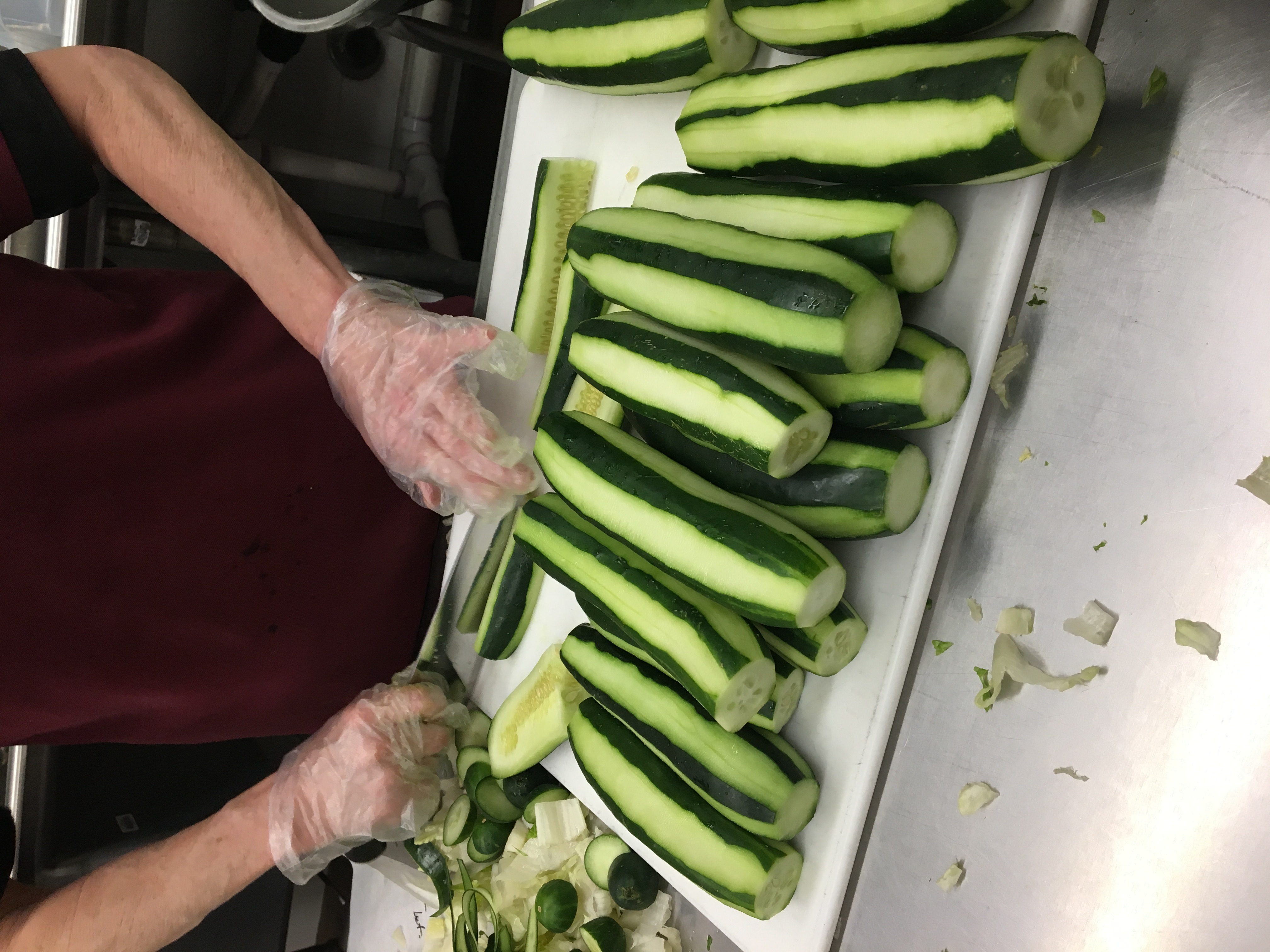 A person wearing gloves is slicing cucumbers on a cutting board, with several whole cucumbers arranged neatly in front of them.