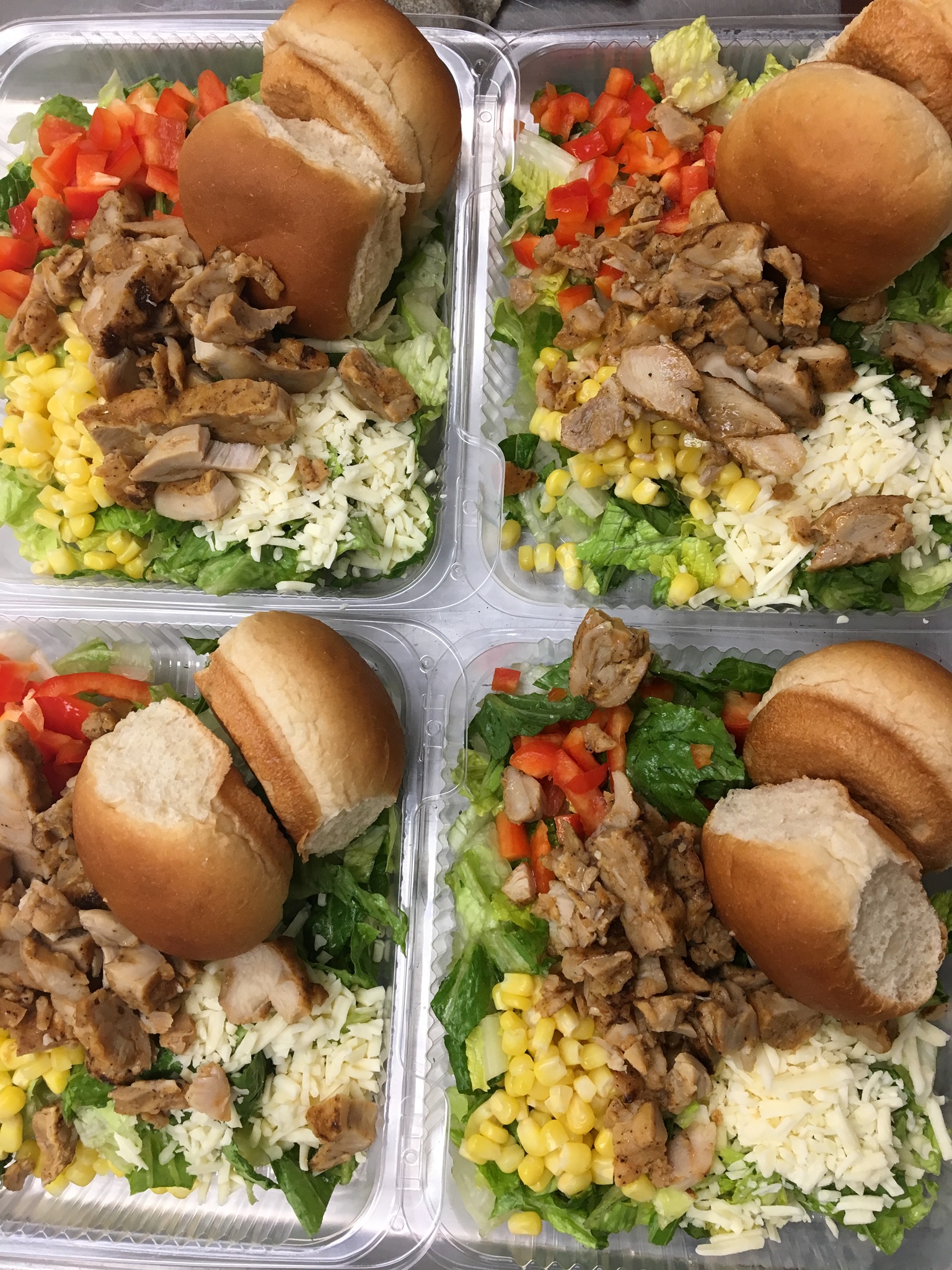 Four plastic containers filled with salads, each containing chopped lettuce, diced tomatoes, corn, shredded cheese, and pieces of grilled chicken, accompanied by two small bread rolls on top.