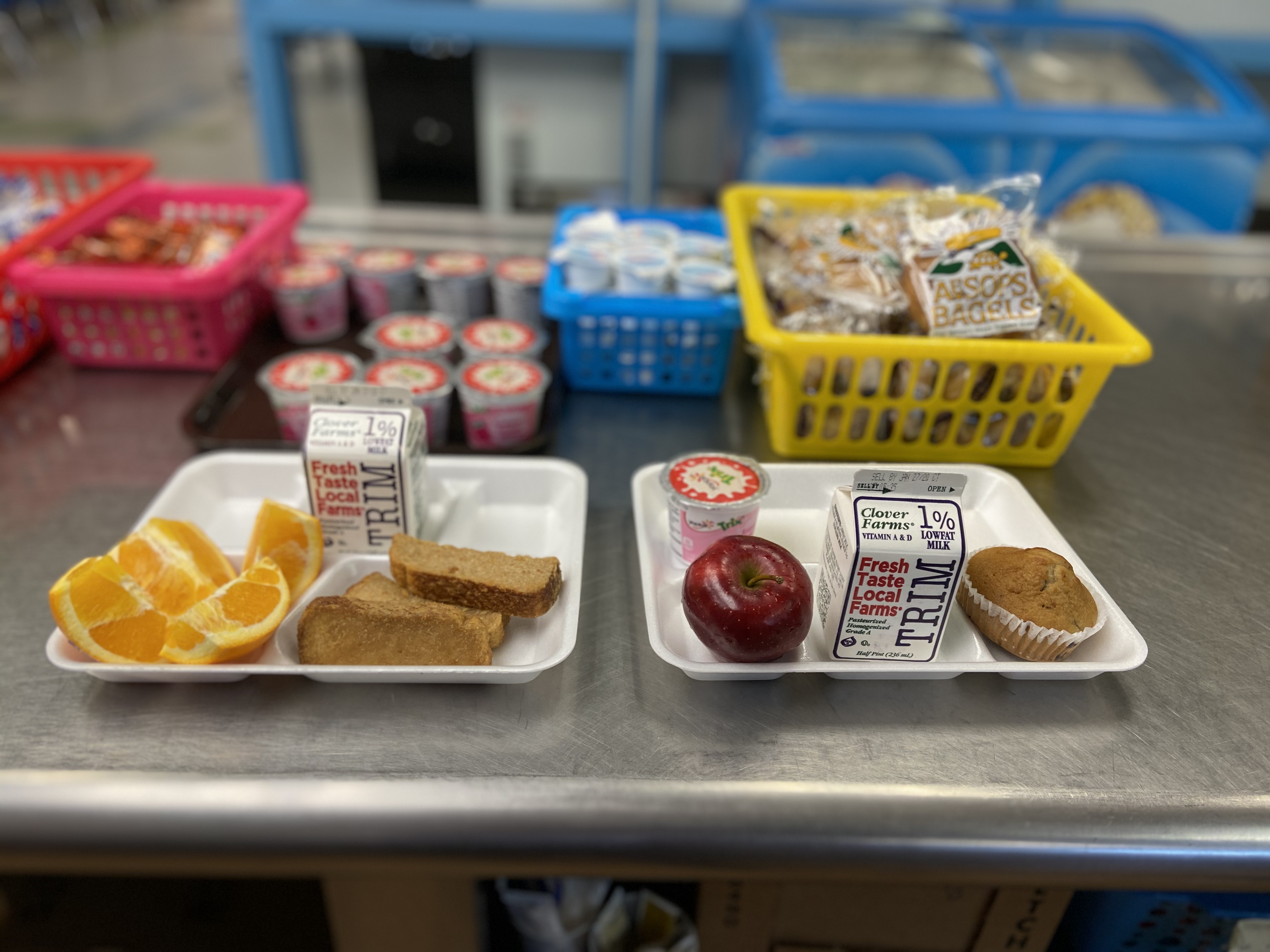 Two lunch trays are displayed on a cafeteria table, one containing orange slices, two granola bars, and milk cartons, while the other has an apple, a muffin, and milk cartons, with colorful baskets of snacks in the background.