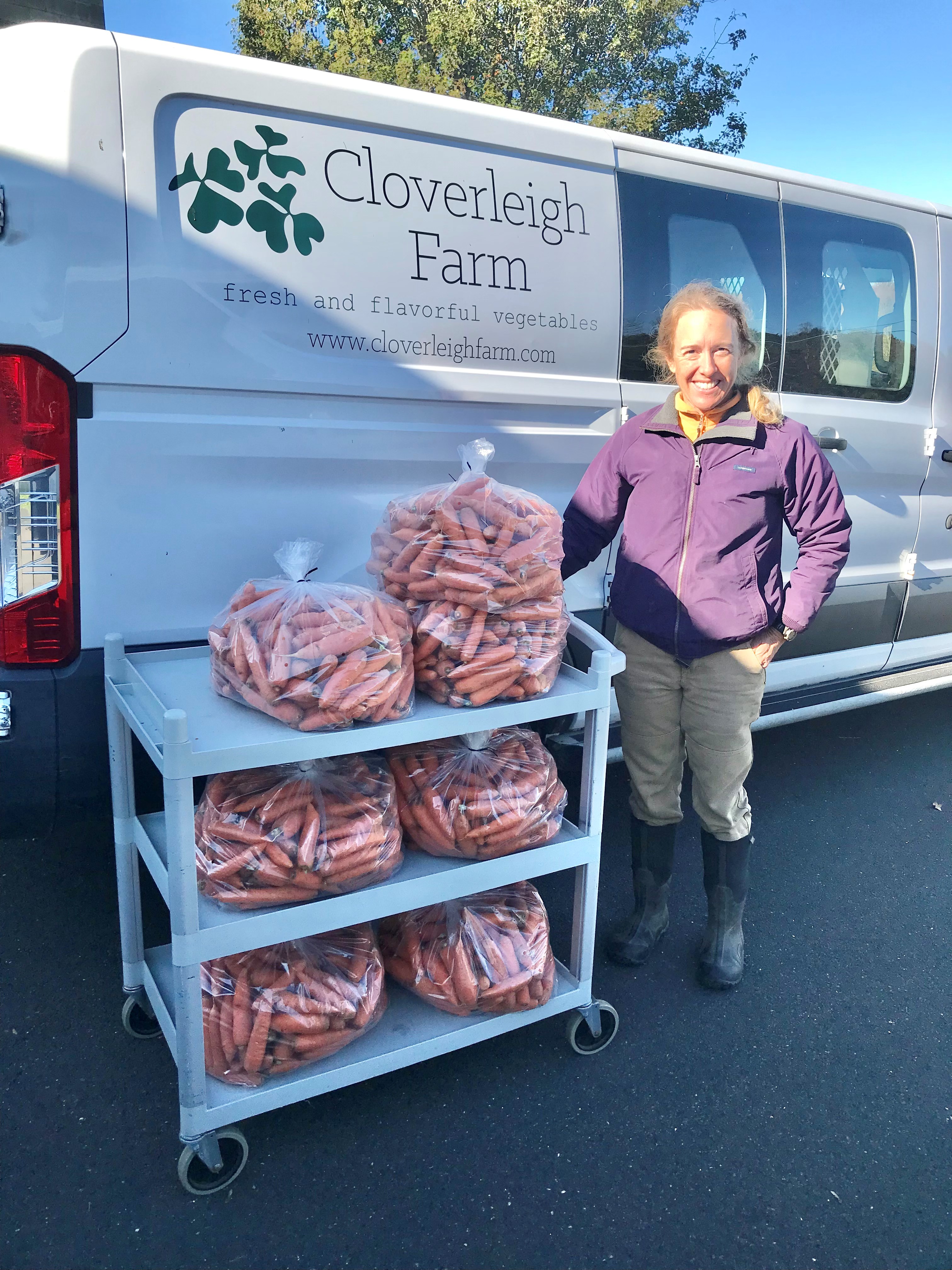 A woman stands next to a cart filled with bags of carrots, in front of a van with the logo of Cloverleigh Farm.