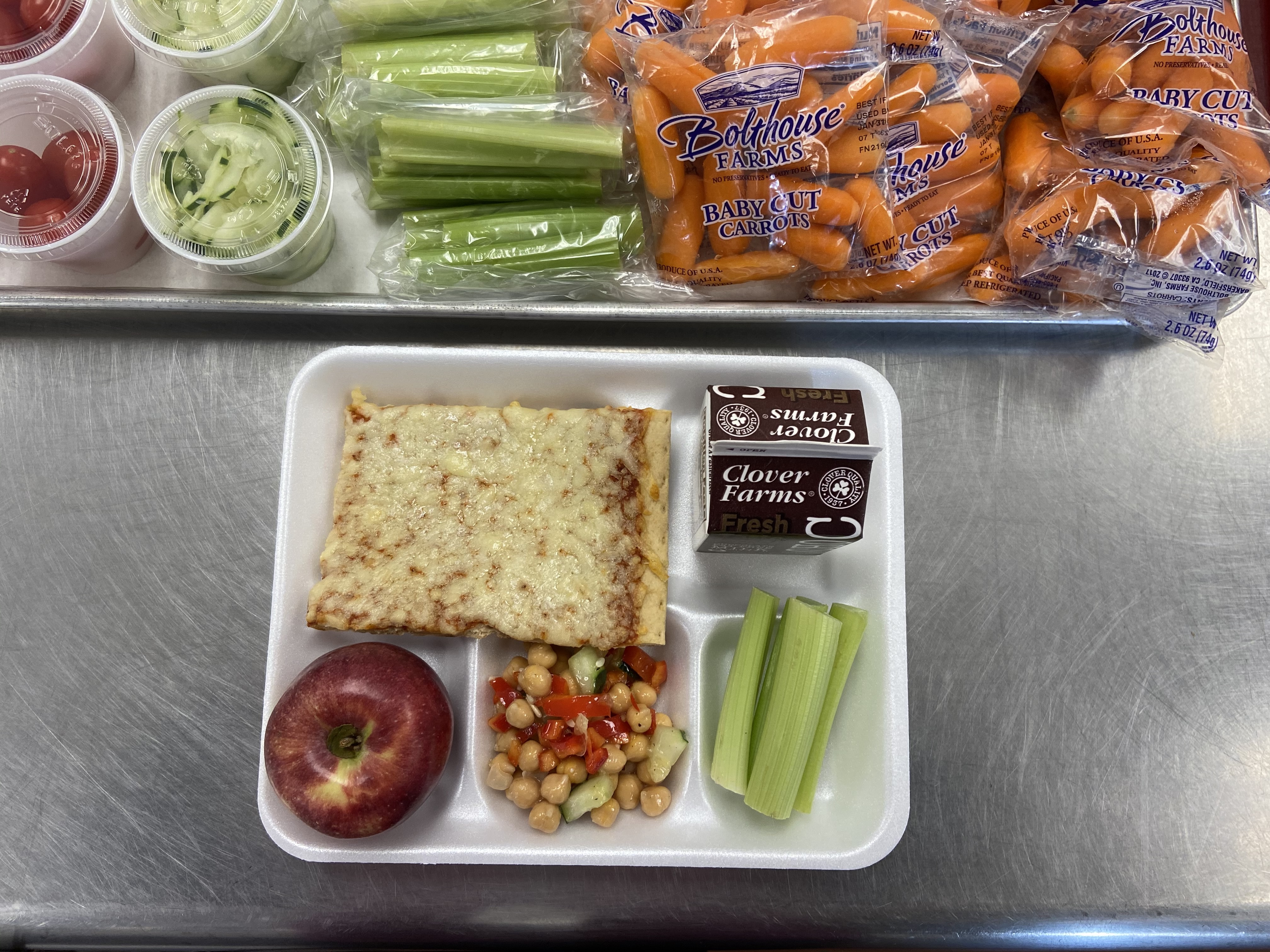 A school lunch tray featuring an apple, a slice of bread, a vegetable salad, two celery sticks, a chocolate pudding cup, and a side of baby carrots and celery sticks in containers.
