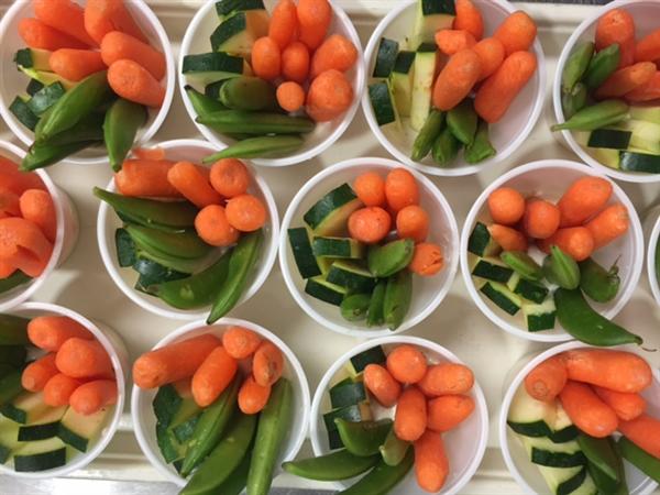 A top-down view of multiple cups filled with fresh vegetables, including orange baby carrots, green snap peas, and pieces of cucumber.