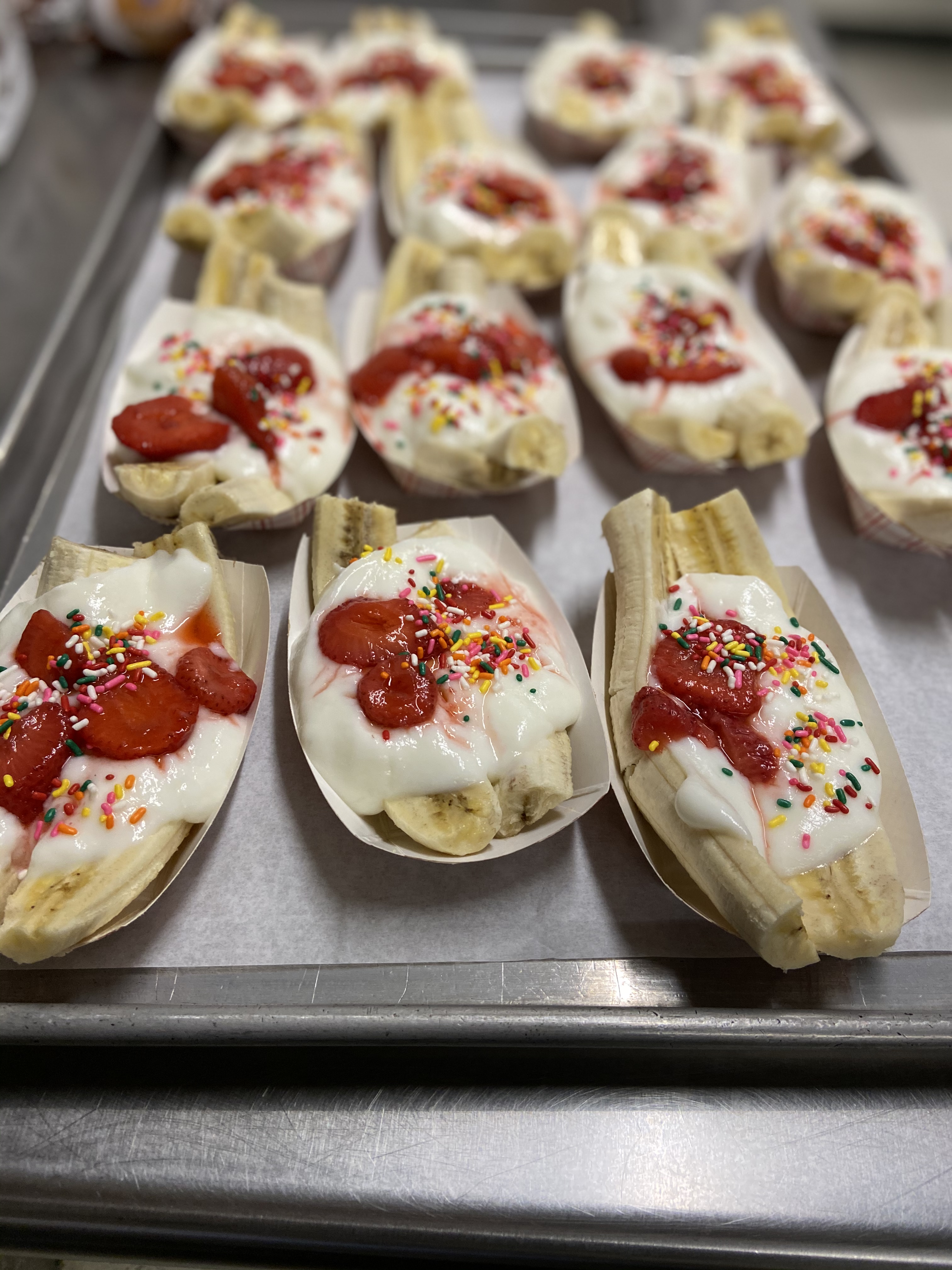 A tray of dessert servings featuring banana splits topped with whipped cream, strawberries, and colorful sprinkles, arranged in paper boats.