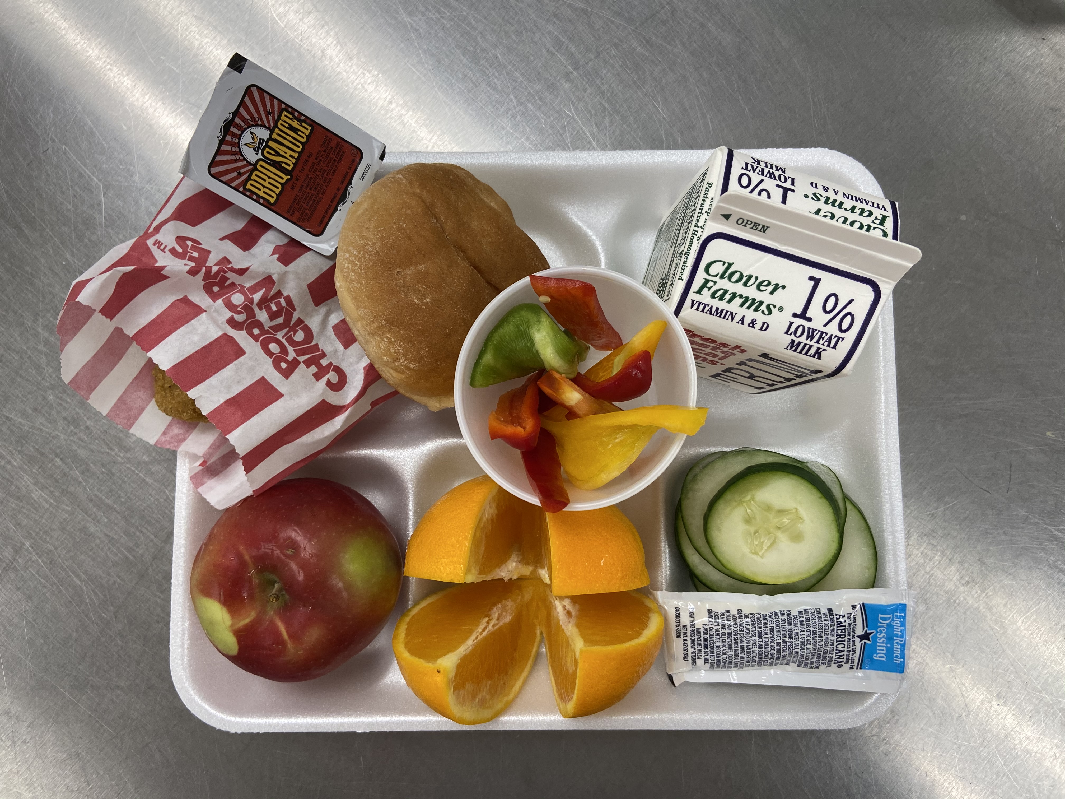 A school lunch tray featuring an apple, orange slices, cucumber slices, a small cup of mixed vegetables, a hamburger bun, a carton of milk, and a small container of dipping sauce.