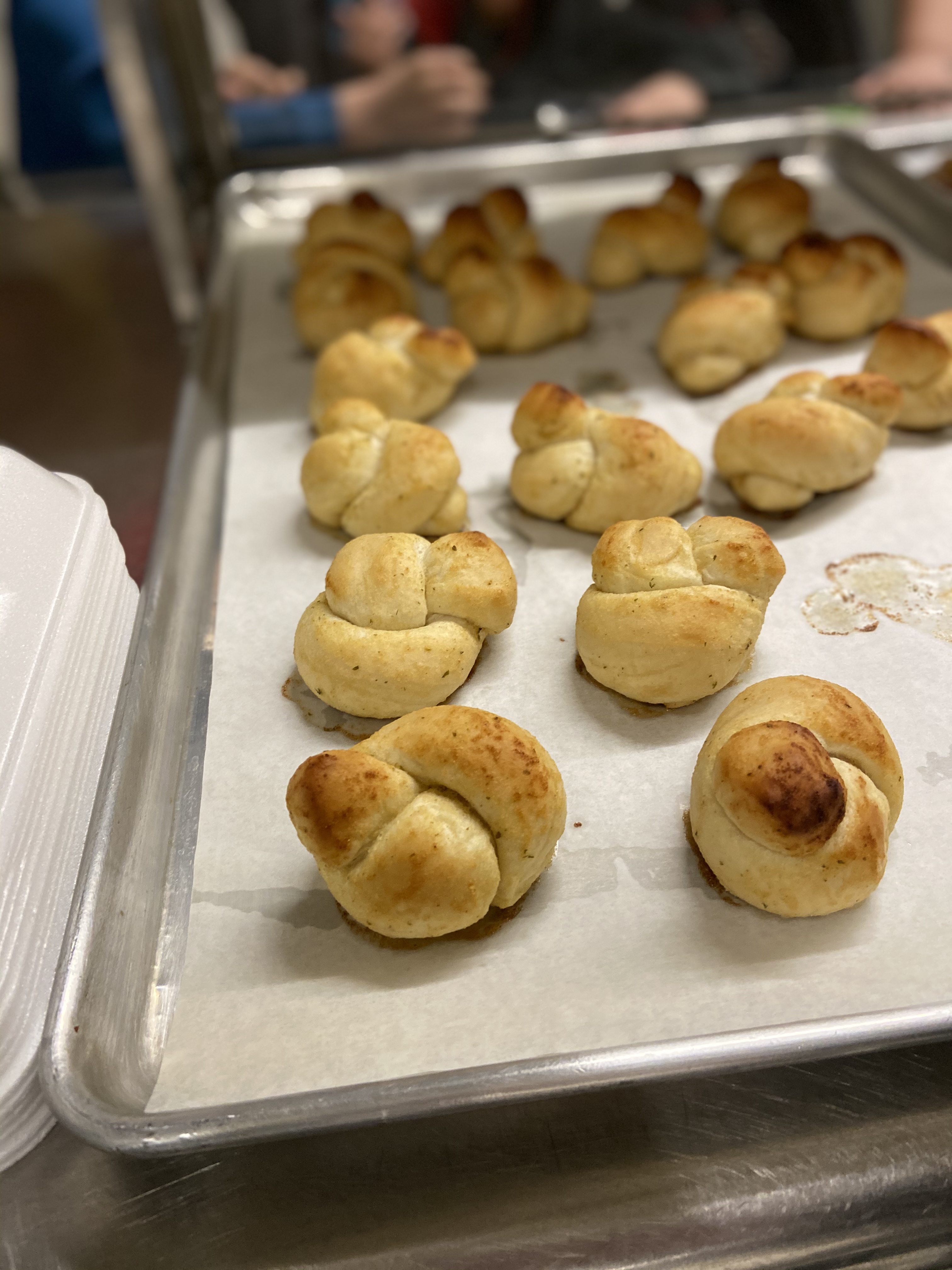 A tray of freshly baked, golden-brown dinner rolls arranged neatly, with a blurred background of people.