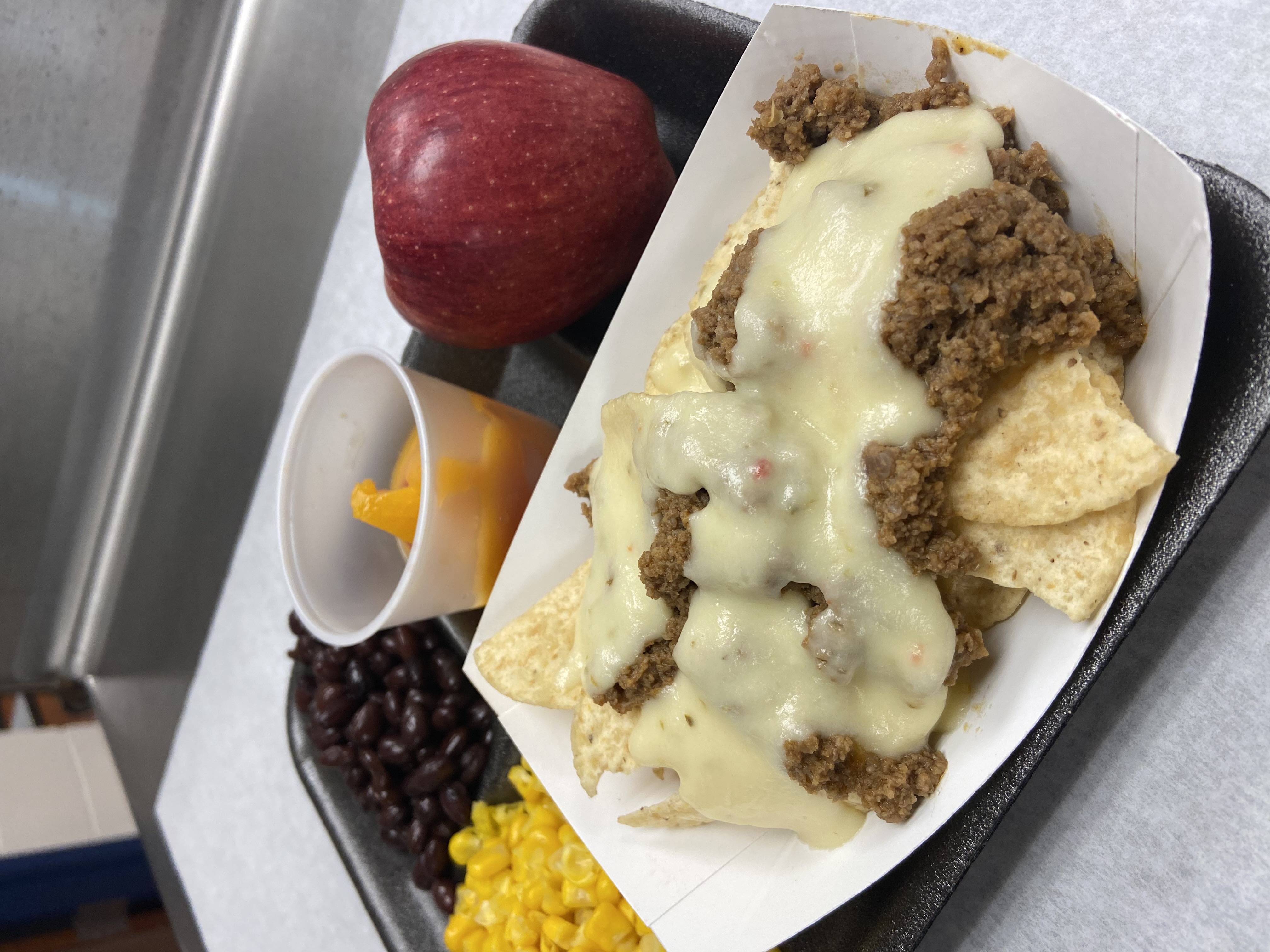 A cafeteria tray featuring nachos topped with ground beef and melted cheese, a cup of fruit, black beans, corn, and a red apple.