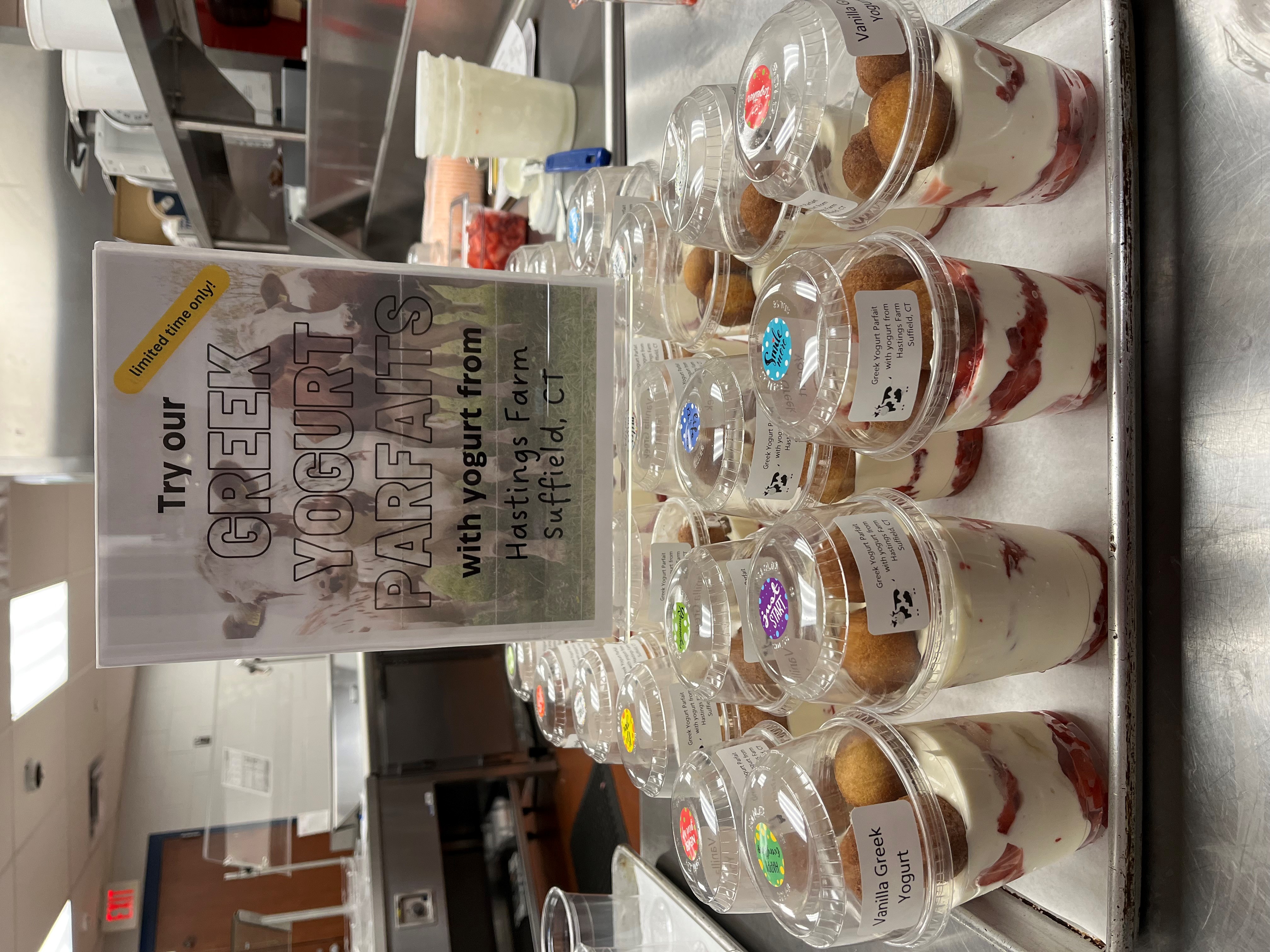 A display of Greek yogurt parfaits in clear cups, topped with granola and fruit, arranged on a tray in a kitchen setting, with a sign promoting the parfaits in the background.