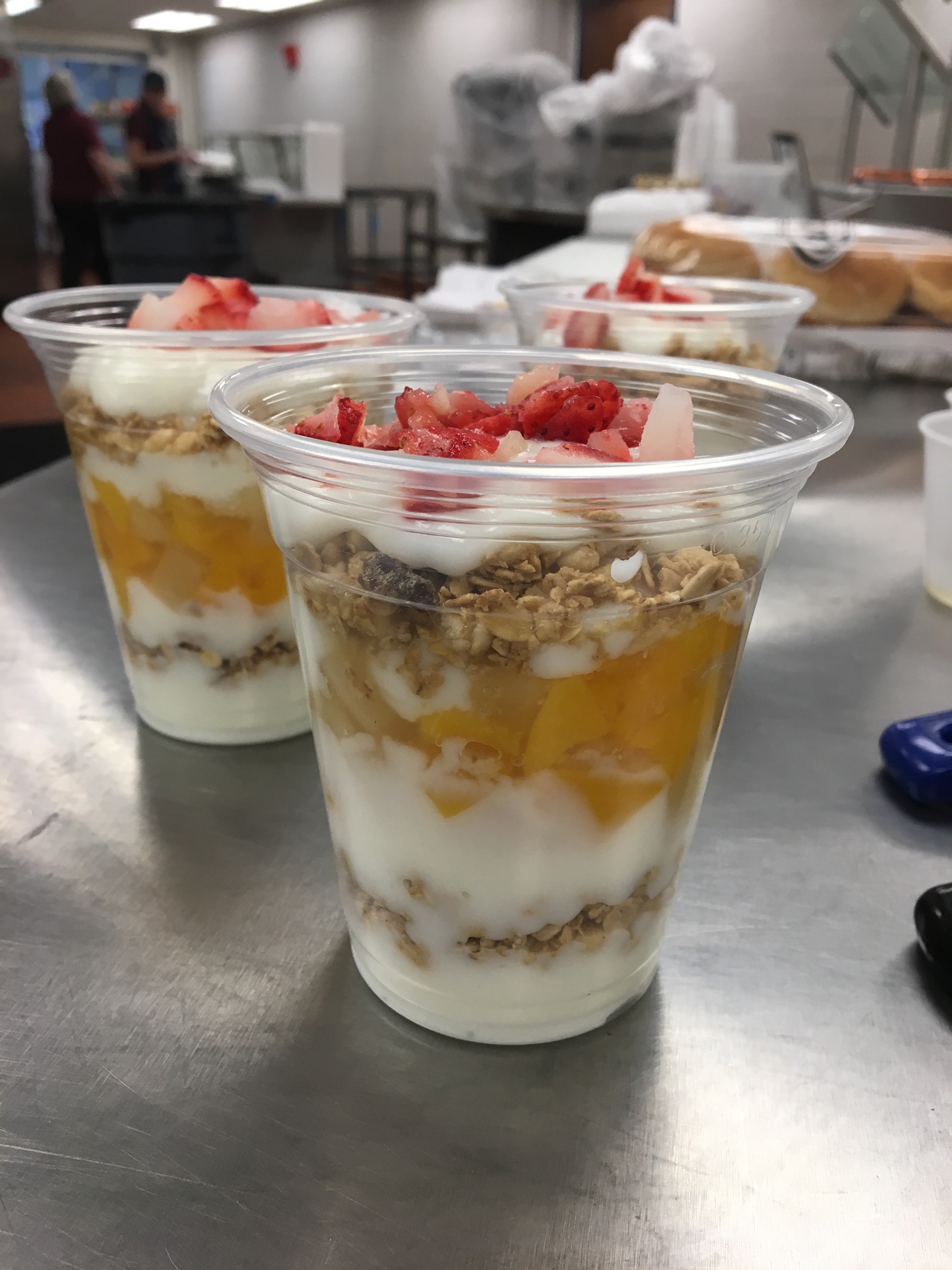 A clear plastic cup filled with layers of yogurt, granola, and fruit, topped with diced strawberries, placed on a table with other food items in the background.