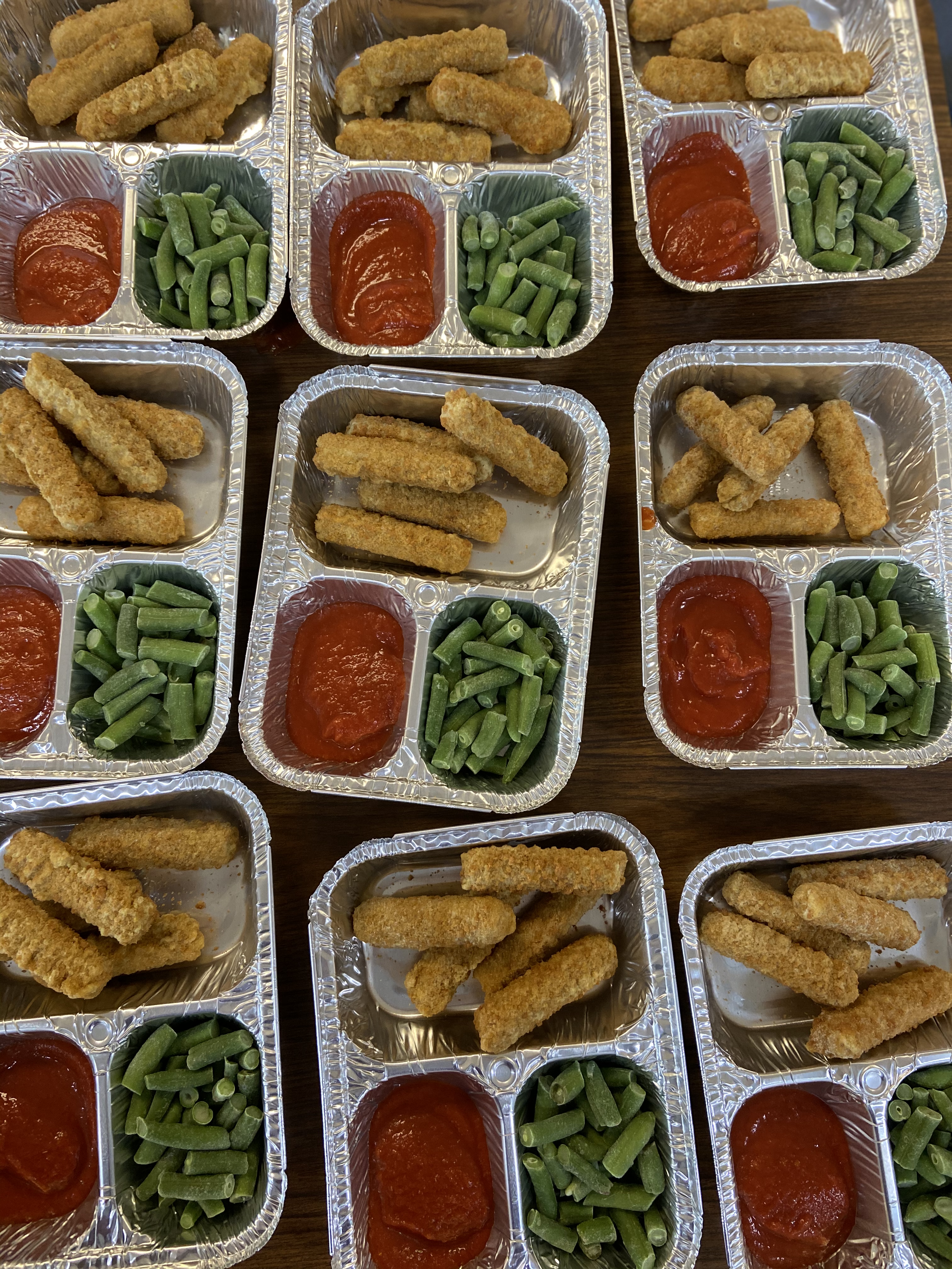 A top-down view of multiple aluminum food trays, each containing breaded chicken strips, green beans, and small cups of marinara sauce.
