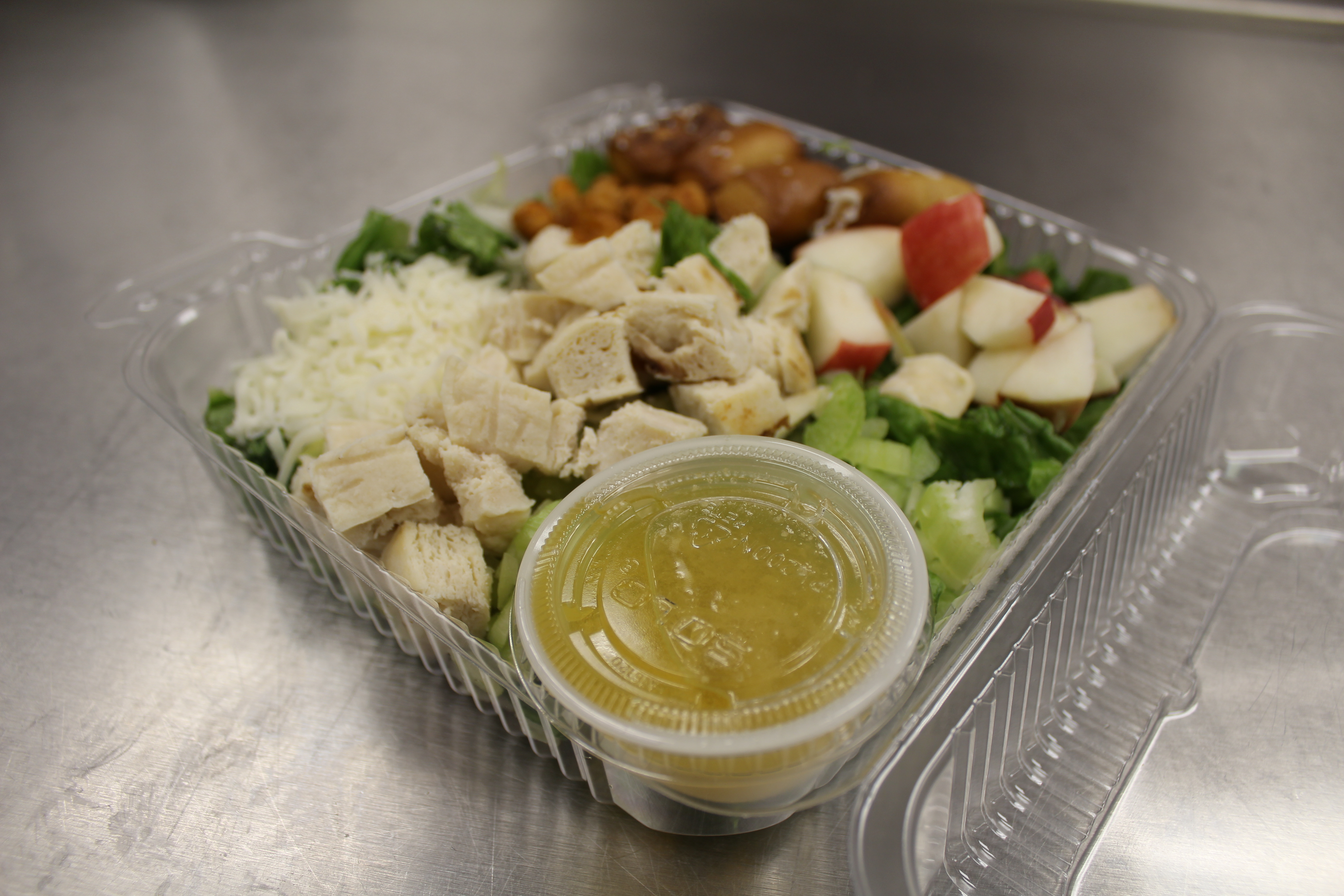 A clear plastic container filled with a salad featuring chopped lettuce, diced chicken, shredded cheese, apple pieces, and a small cup of dressing on the side.