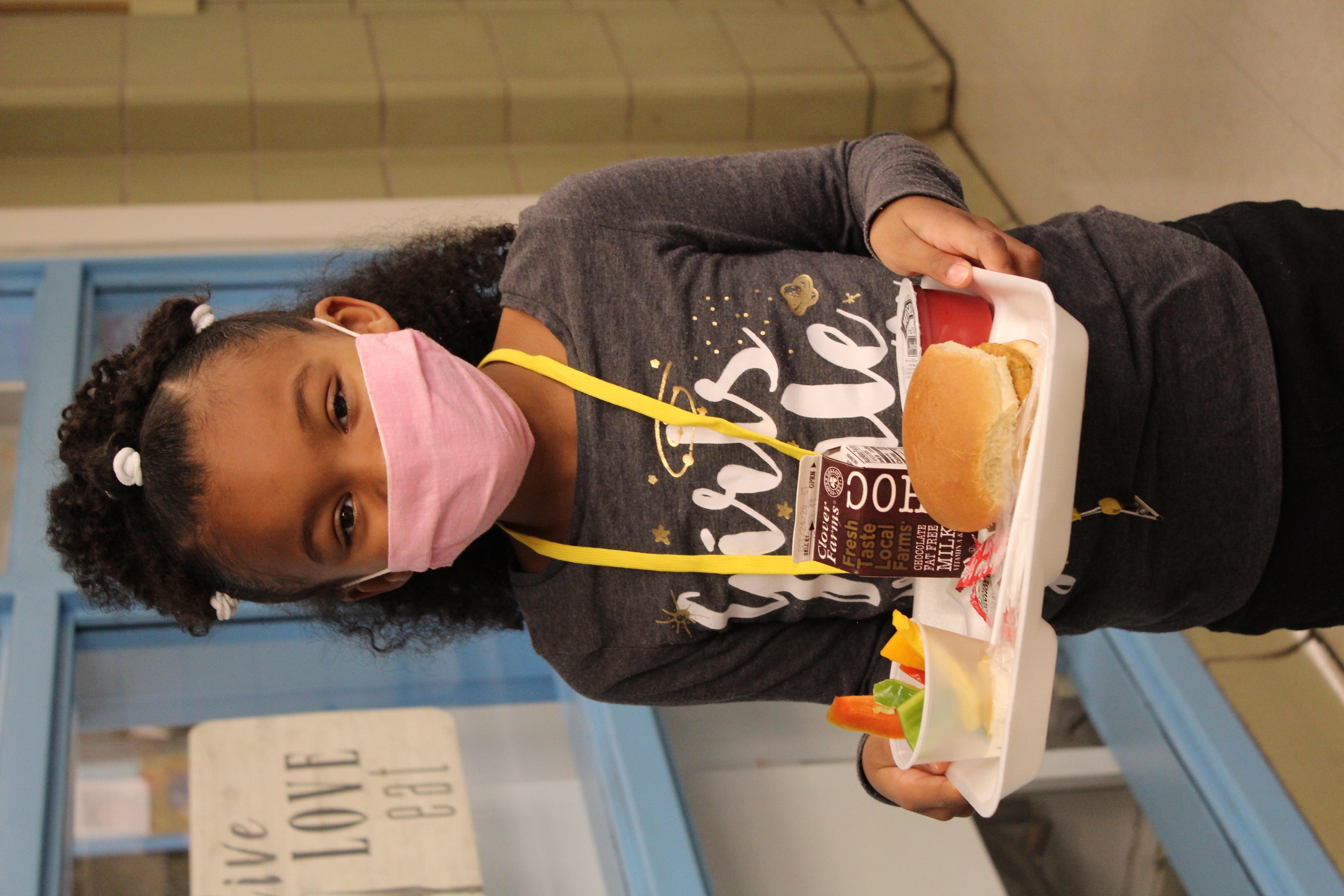 A young girl wearing a pink mask and a gray shirt with the text 'Girls Rule' smiles while holding a lunch tray filled with a sandwich, fruit, and a drink.