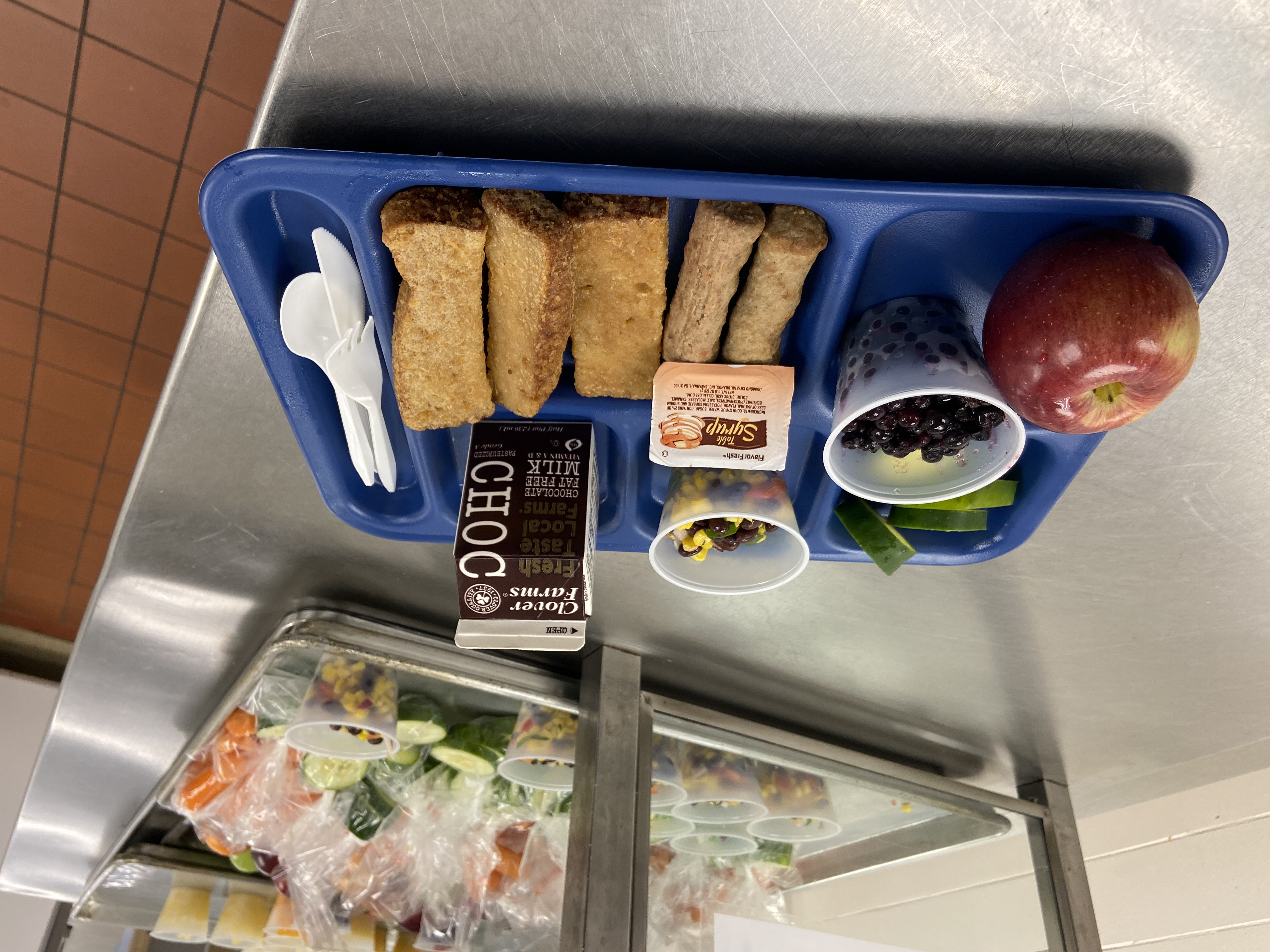 A blue cafeteria tray holds an apple, a cup of mixed fruit, a carton of chocolate milk, a small container of dipping sauce, and several rolled-up snacks, with plastic utensils beside them.