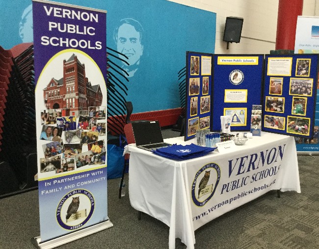 A display booth for Vernon Public Schools featuring a banner, a table with promotional materials, and a backdrop with images related to the school district.