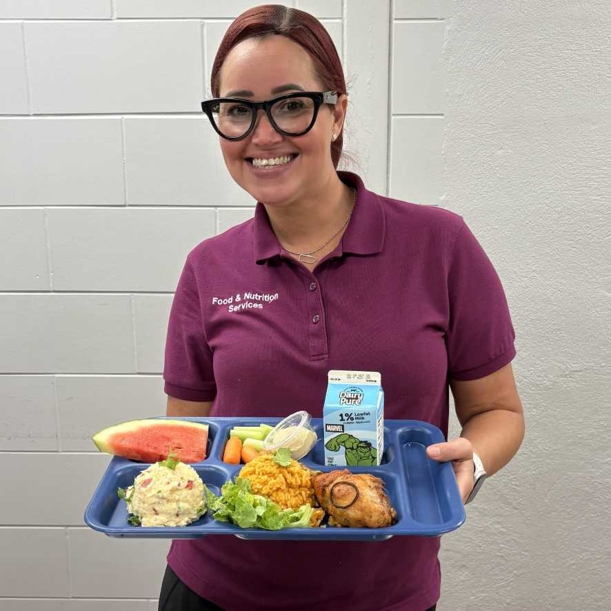 Two women in black aprons and masks stand in a kitchen holding large heads of cabbage, with cooking equipment visible in the background.