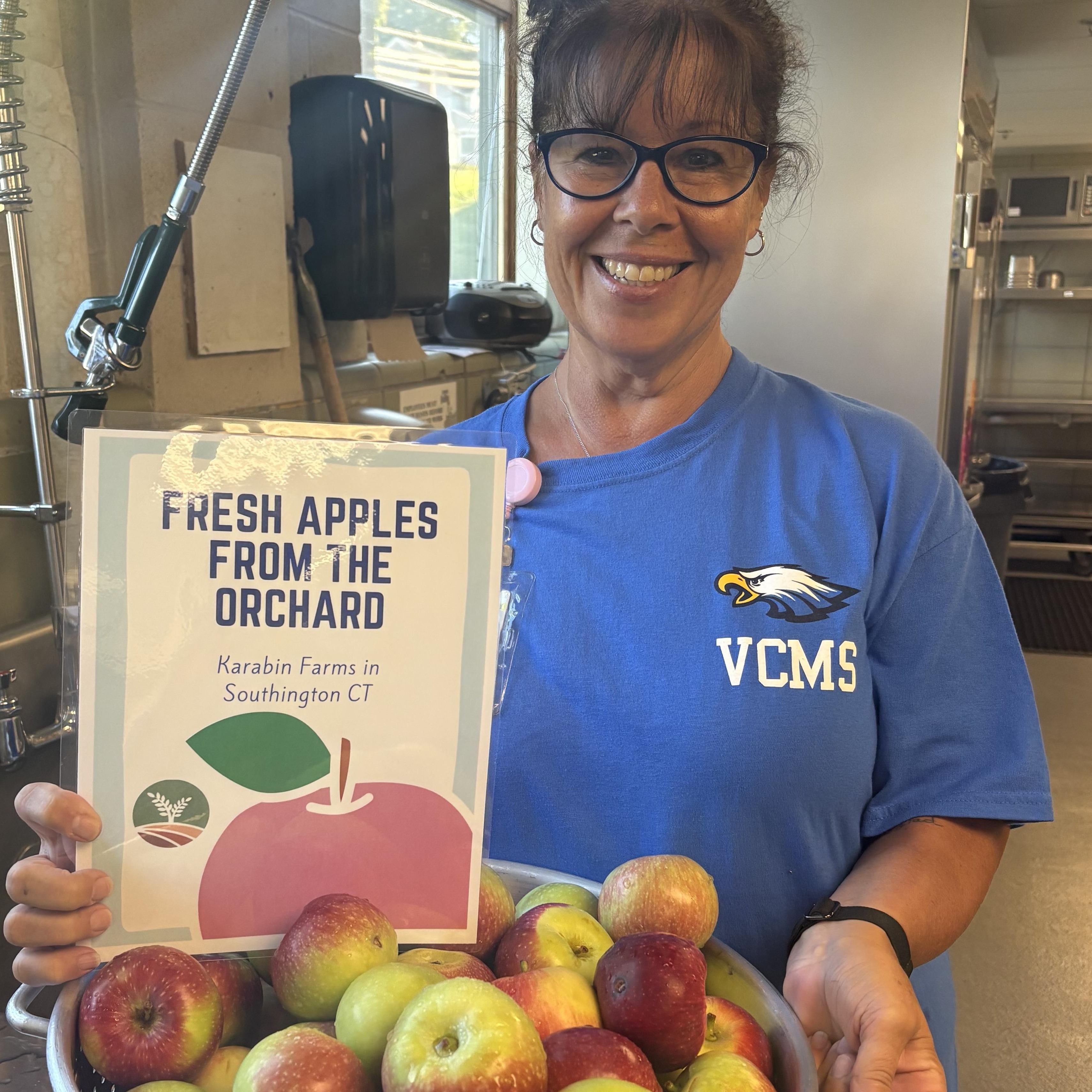 Two women wearing black aprons and caps stand together in a kitchen, holding a large glass jug with a label, smiling at the camera.