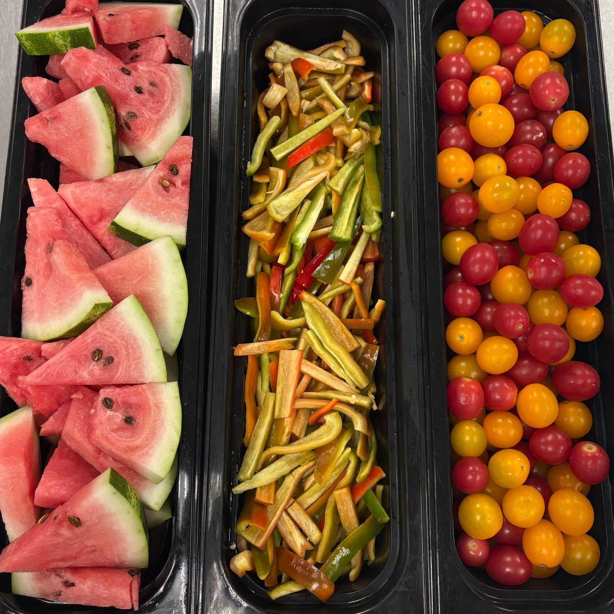 Two lunch trays are displayed on a cafeteria table, one containing orange slices, two granola bars, and milk cartons, while the other has an apple, a muffin, and milk cartons, with colorful baskets of snacks in the background.