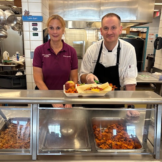A woman wearing a colorful mask smiles while holding a tray filled with numerous cups of dessert topped with whipped cream and fruit, set in a kitchen environment.