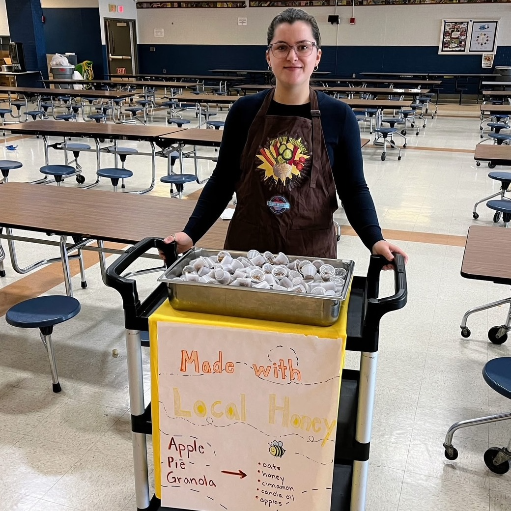 A school lunch tray featuring an apple, orange slices, cucumber slices, a small cup of mixed vegetables, a hamburger bun, a carton of milk, and a small container of dipping sauce.