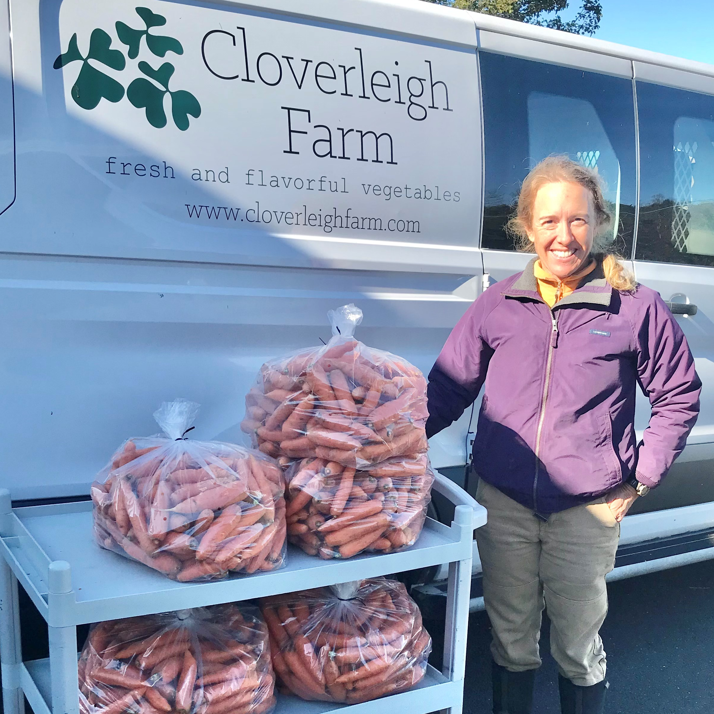 A woman stands next to a cart filled with bags of carrots, in front of a van with the logo of Cloverleigh Farm.
