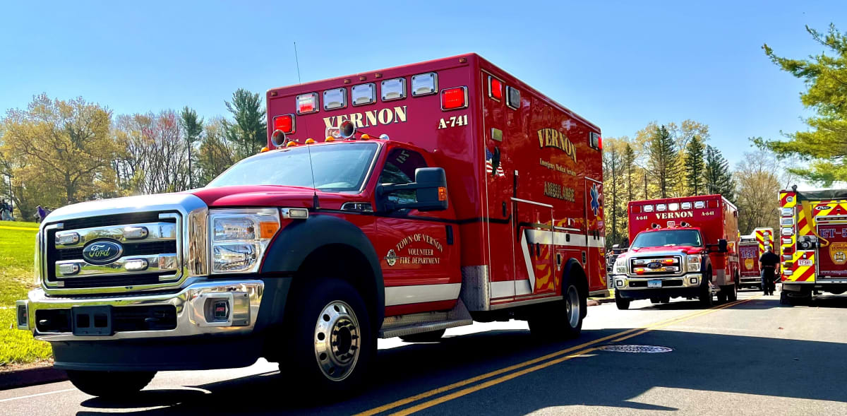 A red emergency ambulance is parked on a sunny street, with additional emergency vehicles visible in the background.
