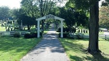 A pathway leading through a cemetery, framed by a white archway and surrounded by trees and gravestones.