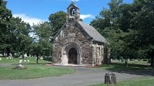 A stone chapel with a bell tower, surrounded by trees and a grassy area, set within a cemetery.