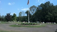 A circular area in a cemetery featuring a flagpole with an American flag, surrounded by gravestones and greenery under a clear blue sky.