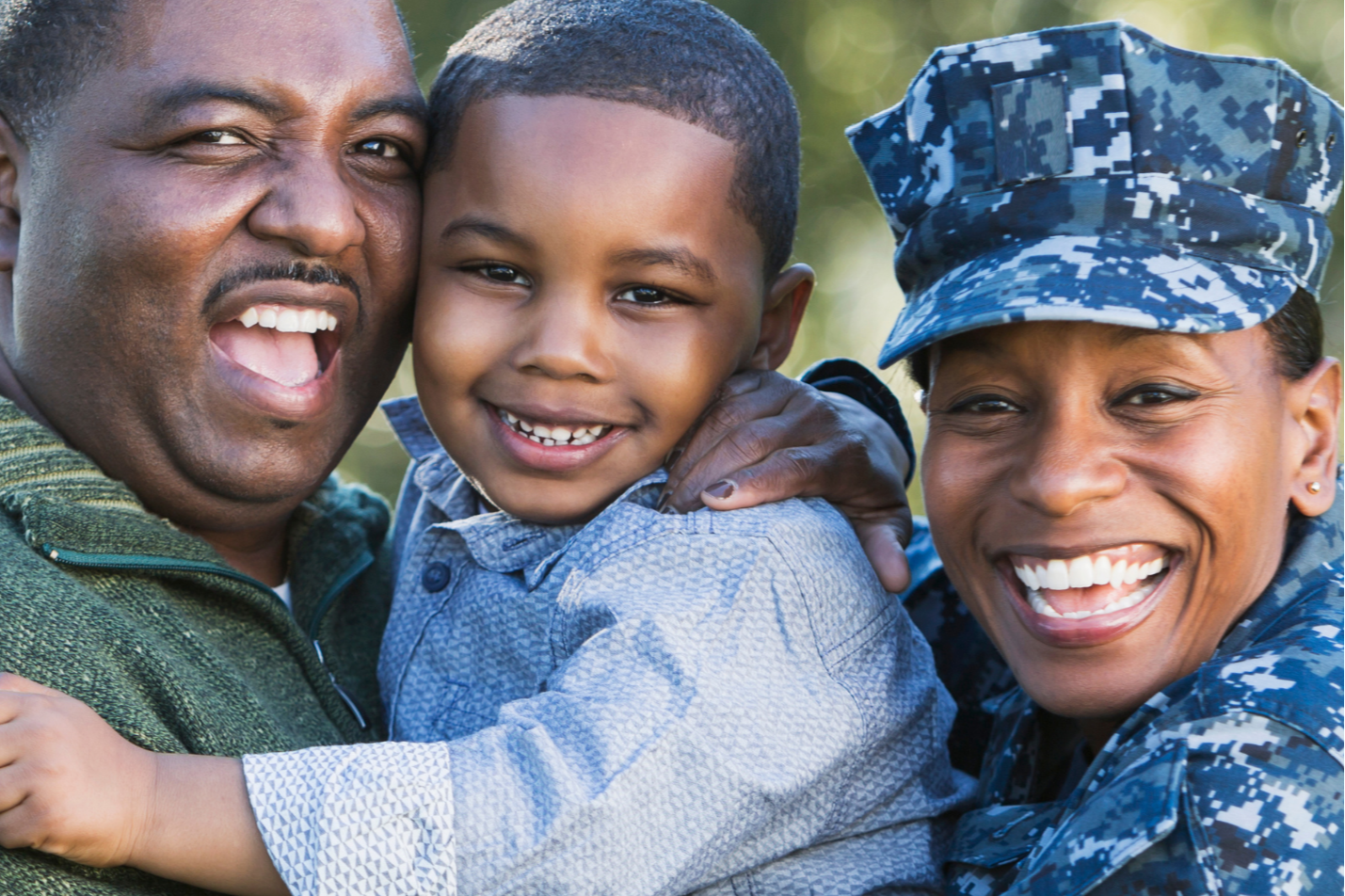 Man in green and woman in military uniform embrace a smiling child. All are smiling. Trees in the background.