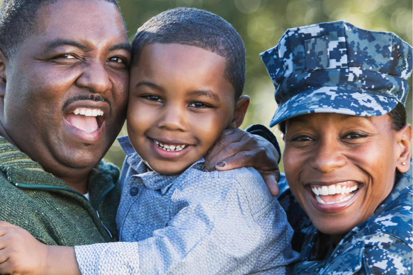 A man and woman, both smiling, embrace a child. The woman is in a camouflage uniform, the man wears a green shirt, and the background is blurred.