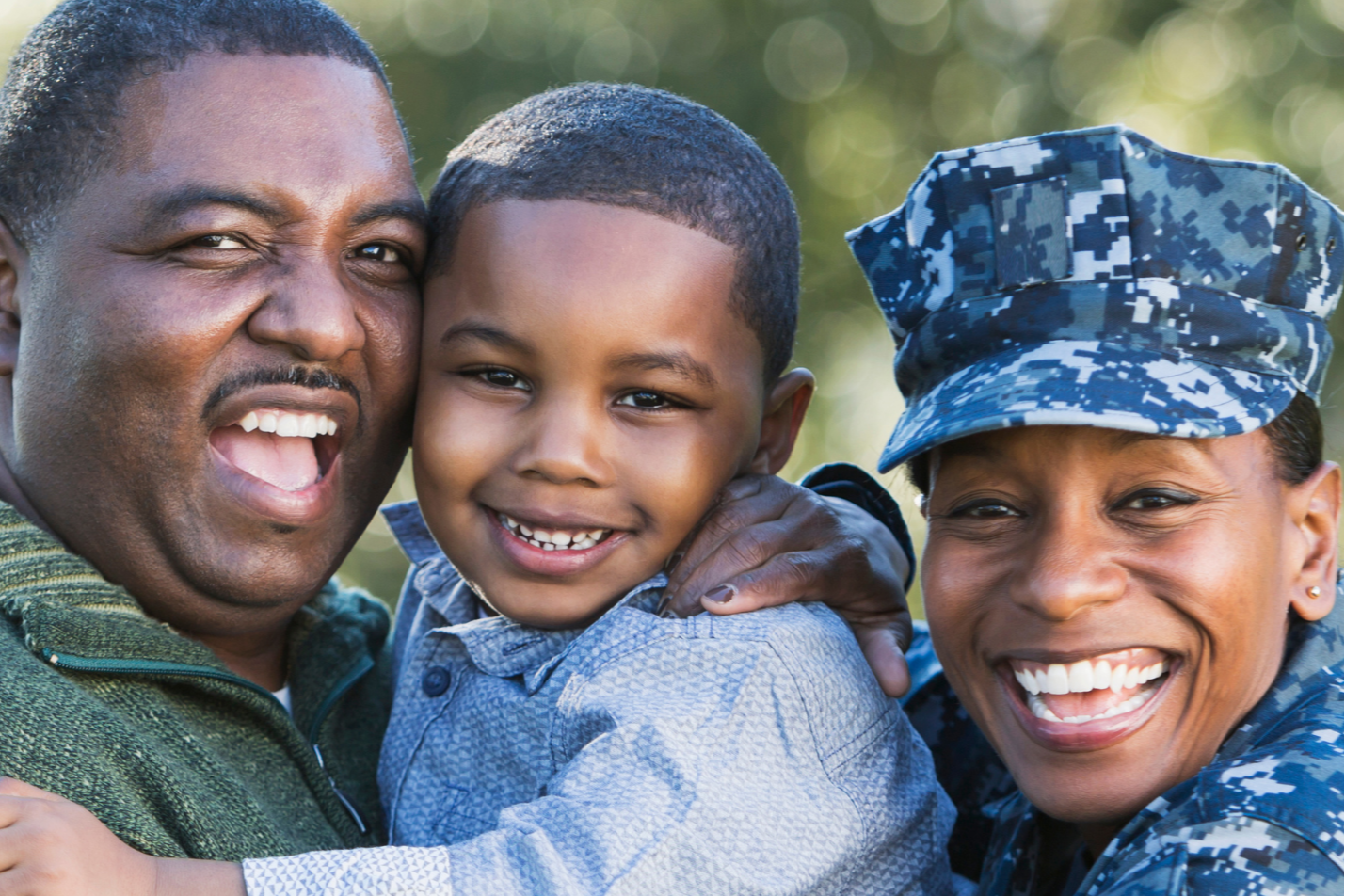 A smiling father, mother in military uniform, and their son all embrace each other in a park.