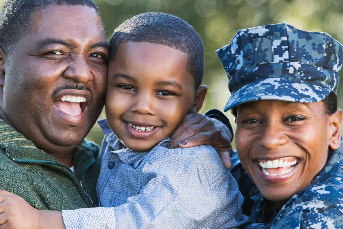 Family of three smiling. Man in green sweater, woman in camouflage, and child in blue shirt. Blurry greenery in background.