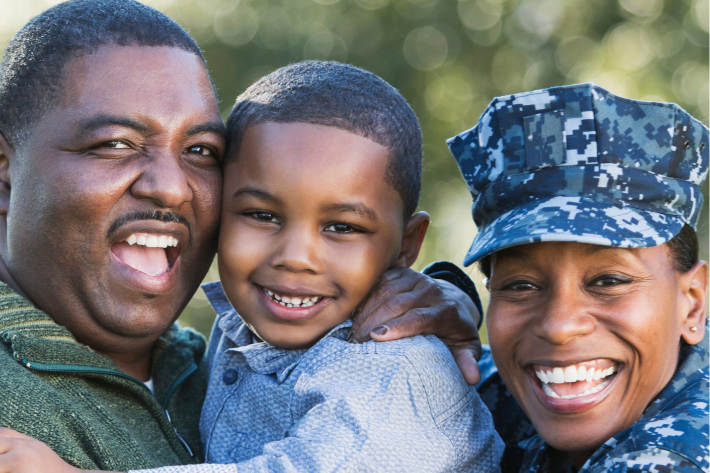 A family outdoors, smiling. A young boy holds his parents' arms. Father in green, mother in camouflage, child in blue. Background is blurred.