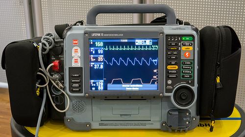 A medical monitoring device with a large screen displaying vital signs and waveforms, surrounded by various buttons and connectors, placed on a yellow surface.