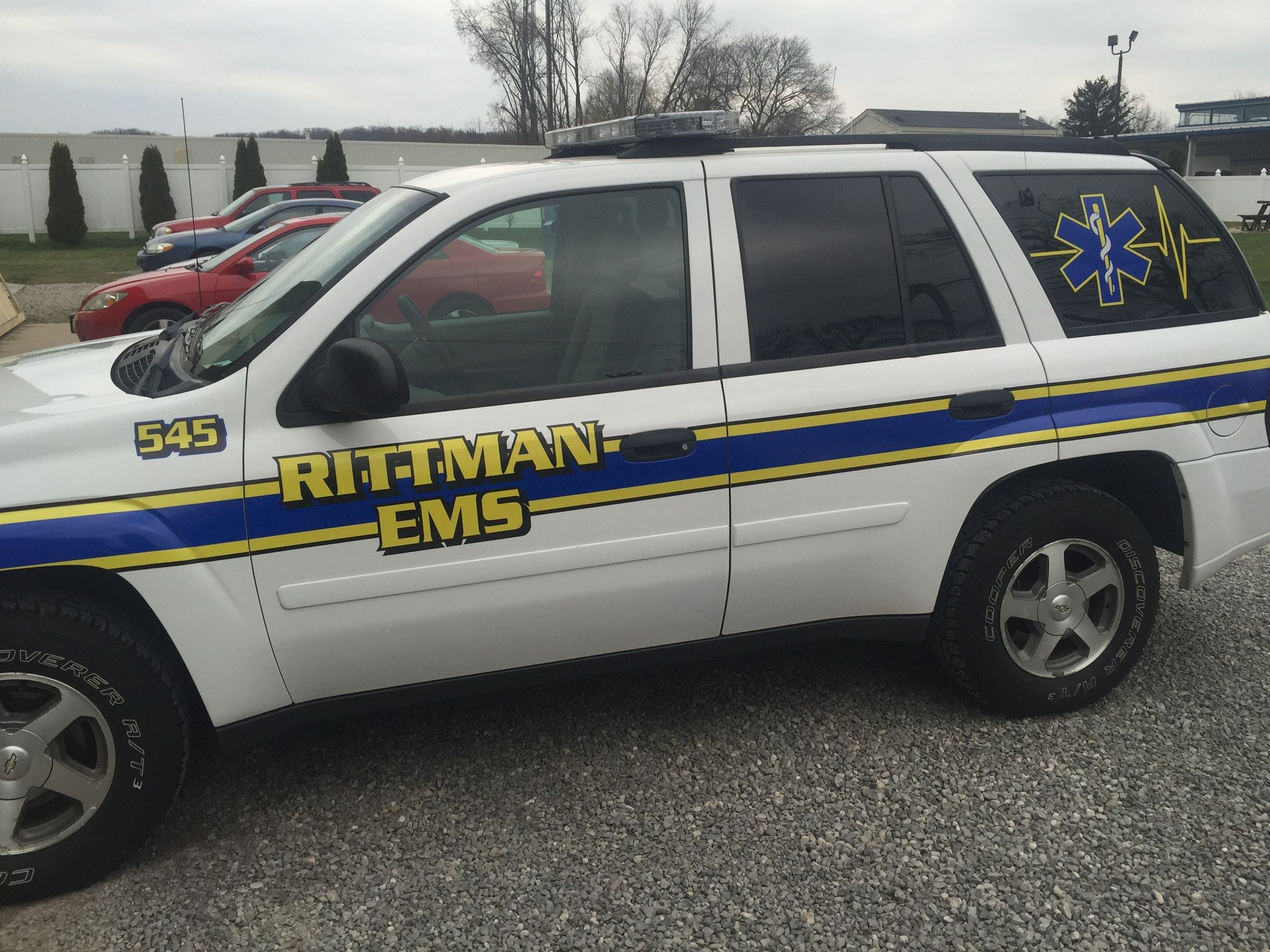 A side view of a white emergency medical services (EMS) vehicle with the words 'Rittman EMS' prominently displayed in yellow and blue lettering, featuring an ambulance symbol on the rear.