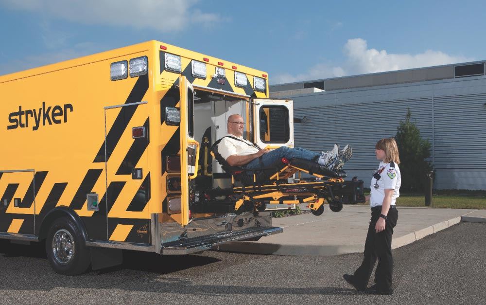 A paramedic assists a patient on a stretcher being loaded into a yellow ambulance marked with 'Stryker,' set against a clear sky and a building in the background.