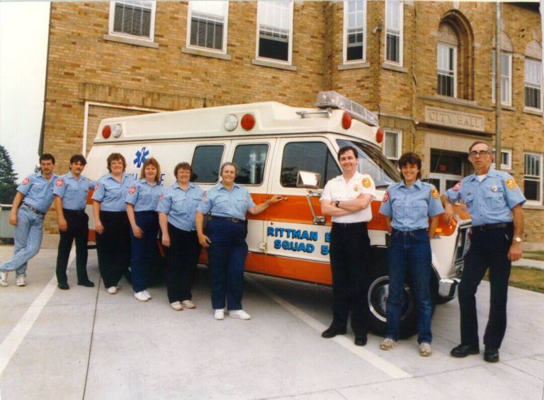 A group of emergency medical personnel stands in front of an ambulance, posing for a photo outside a brick building.