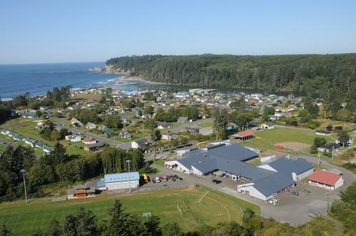 Aerial view of a coastal town with a mix of residential buildings, green fields, and a nearby ocean shoreline surrounded by forested hills.