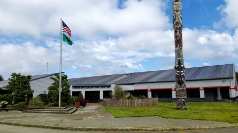 A view of a building with a flagpole displaying the American flag and a green flag, alongside a totem pole, under a partly cloudy sky.