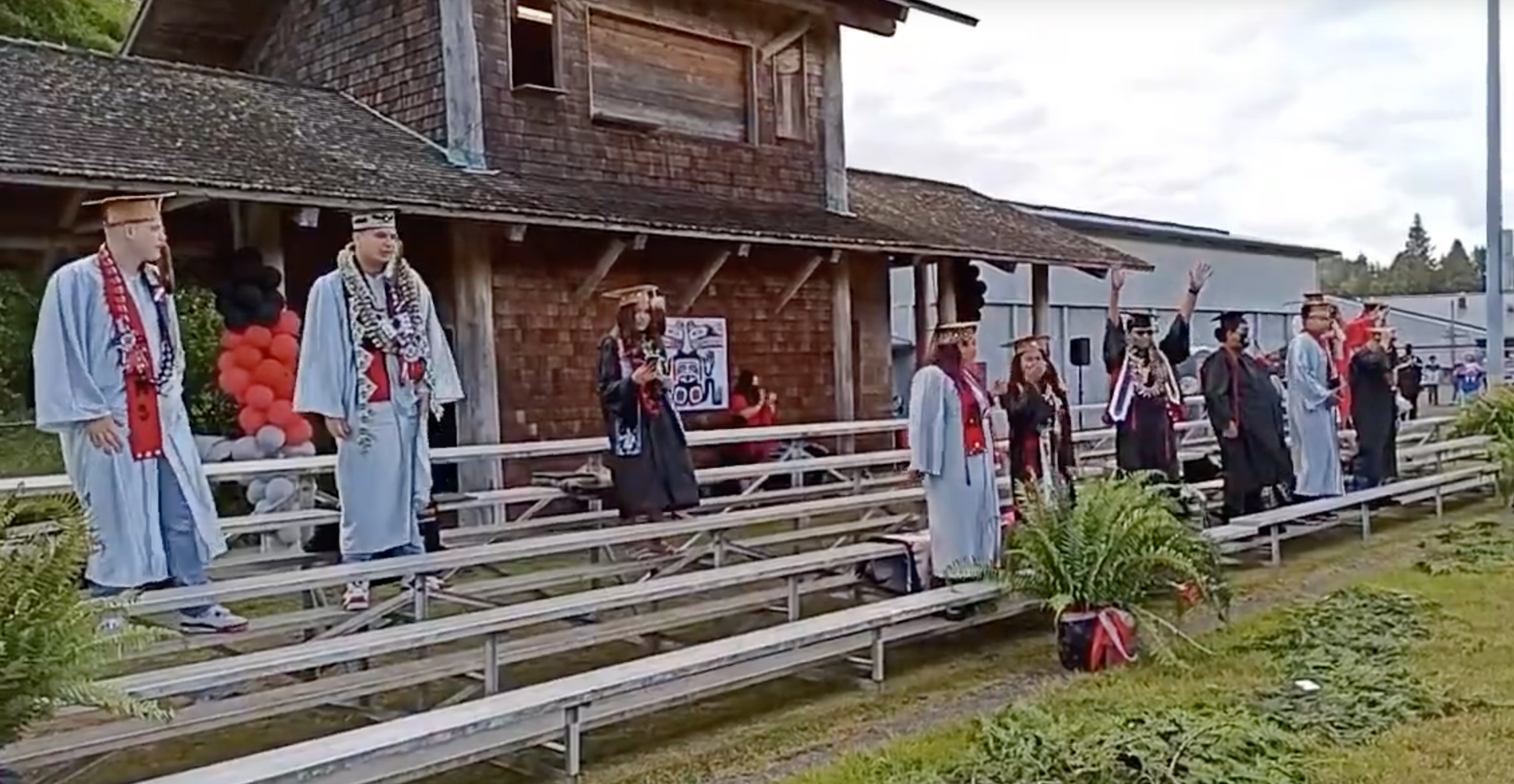 A group of graduates in caps and gowns stands on bleachers outside a building, with decorations in red and black, celebrating their graduation ceremony.