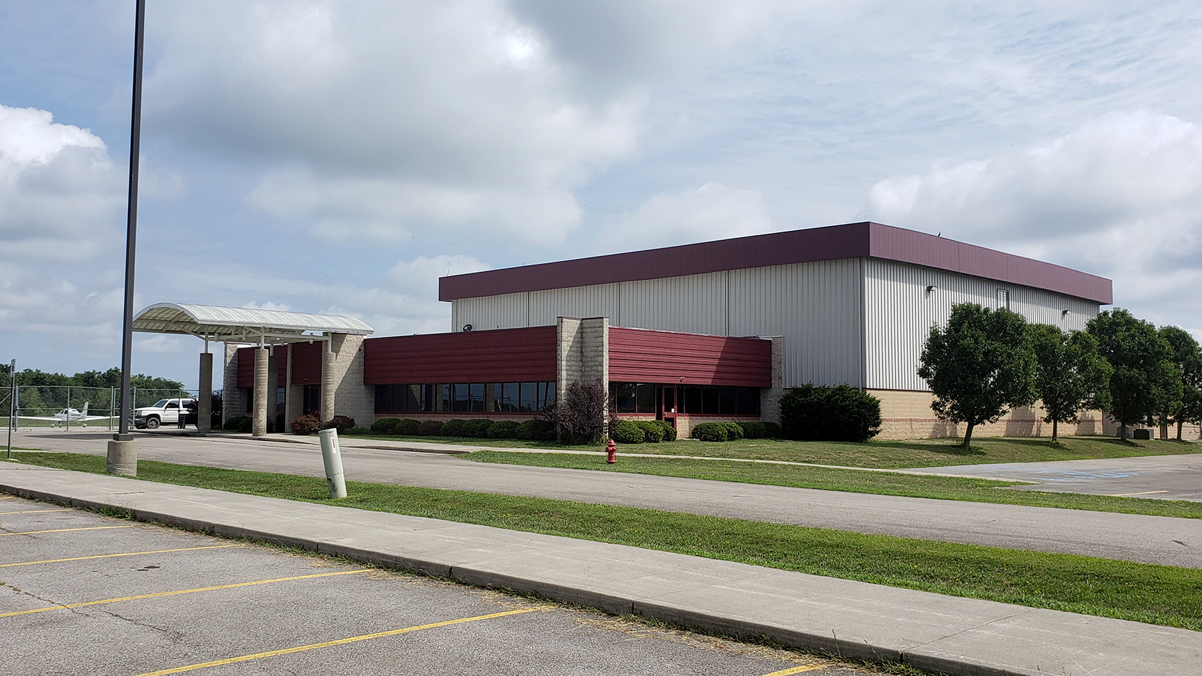 A large, modern building with a maroon roof and a glass entrance, surrounded by green grass and trees under a cloudy sky.