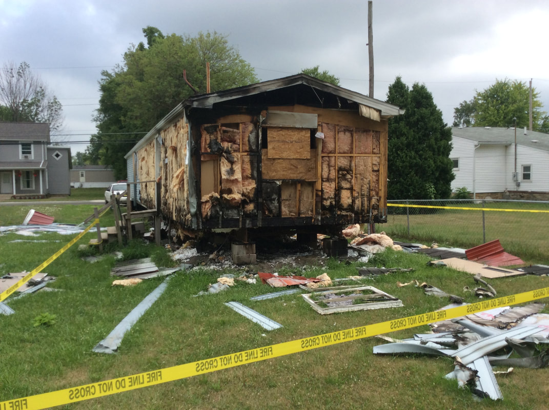 A damaged house with charred and peeling exterior, surrounded by debris and yellow caution tape, indicating a recent fire incident.