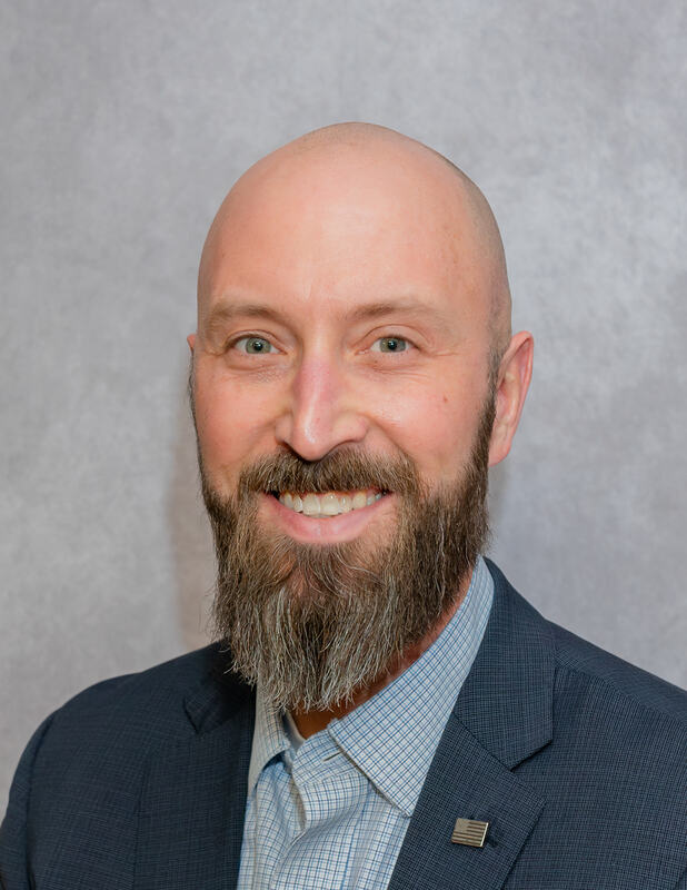 A smiling man with a bald head and a full beard, wearing a dark suit and a light blue checkered shirt, poses against a gray background.