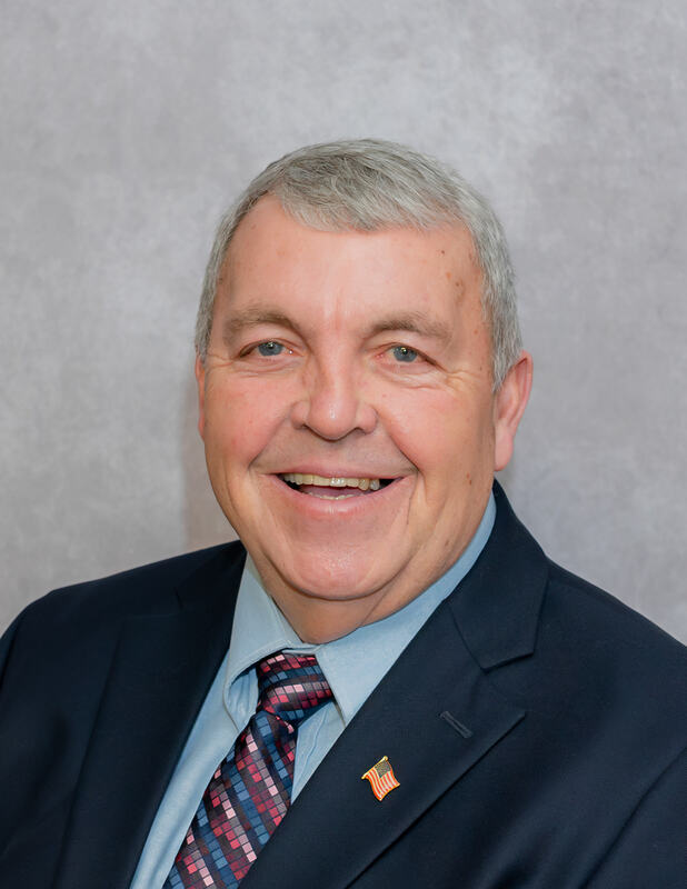 A smiling man with short gray hair, wearing a dark suit and a patterned tie, poses against a light gray background.